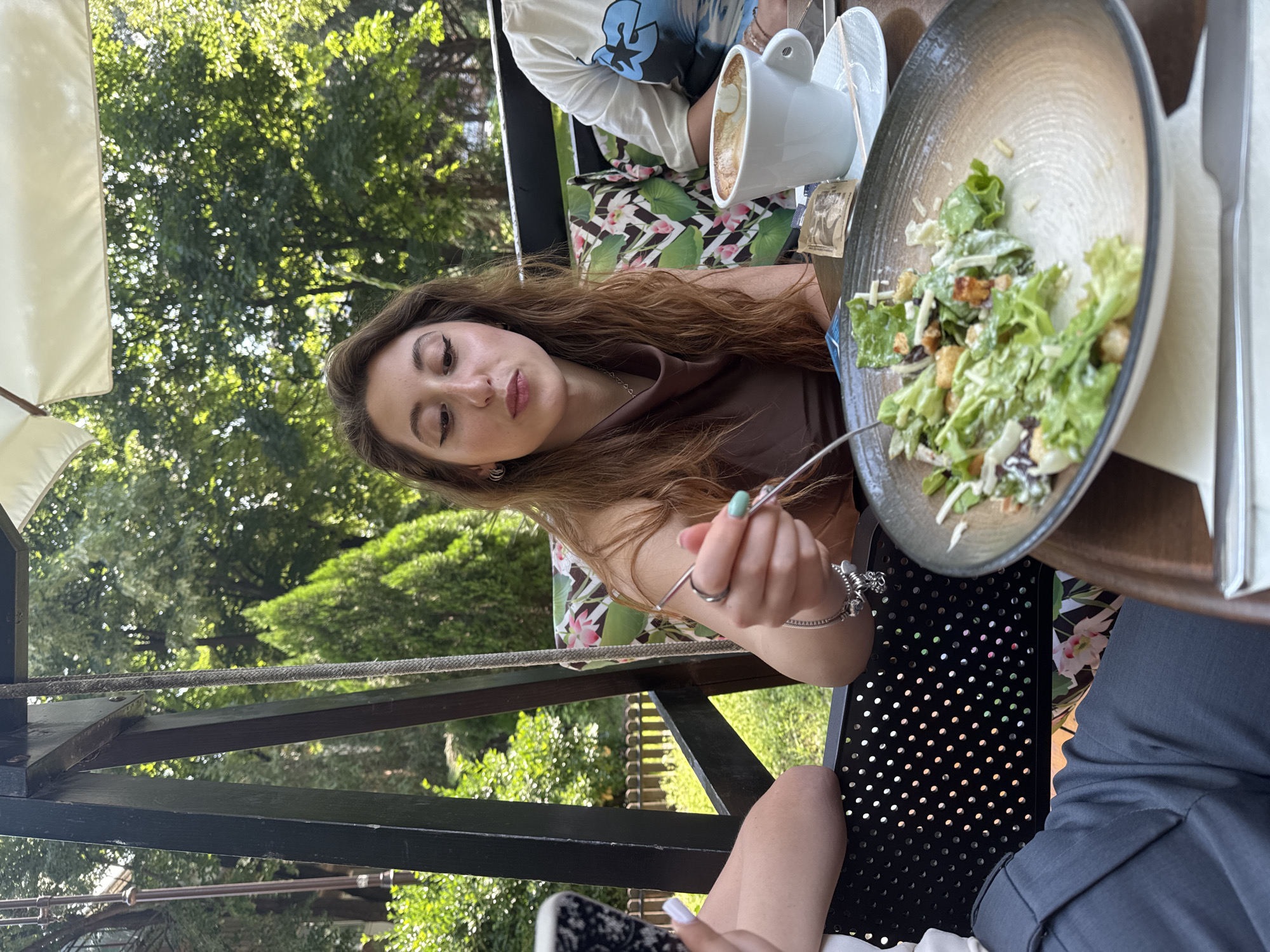 The image depicts an outdoor dining scene on a bright, sunny day, likely at a cafe or garden restaurant. The central subject is a young woman with long, wavy brown hair, wearing a brown, silky-looking top. She is captured from a slightly elevated perspective, looking towards the viewer with a neutral, perhaps pensive, expression. Her right hand, adorned with a silver bracelet and light green nail polish, holds a fork over a salad bowl.
In front of her, a large, dark-rimmed bowl holds a salad primarily composed of green lettuce, croutons, and white cheese or dressing. To the right of the salad, a white mug filled with a beverage resembling a latte or cappuccino, complete with foam art, sits on a saucer.
The scene includes partial views of other individuals. To the upper left, a person wearing a white shirt with blue and black patterns is visible. In the lower right foreground, a leg in grey trousers with white-painted toenails can be seen, indicating another person seated nearby. The table has a dark, possibly wooden surface.
The background is filled with vibrant green trees and foliage, suggesting a lush, natural environment. A white umbrella canopy provides shade on the left side of the frame. A dark, metal support structure, possibly part of an outdoor swing or specialized seating, runs horizontally across the lower middle of the image. The overall atmosphere conveys a relaxed, pleasant outdoor meal. No visible text is present in the image.