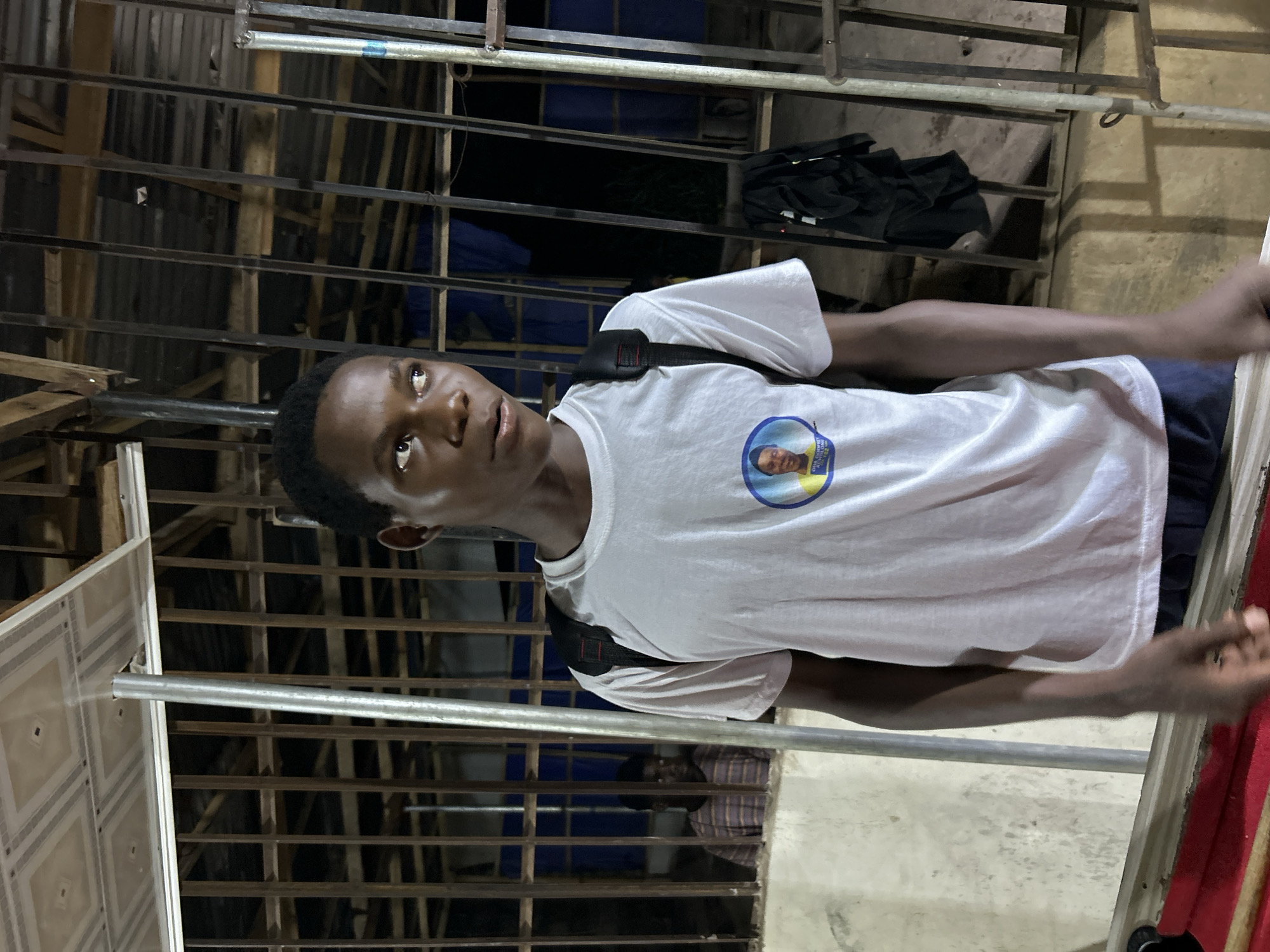 A person wearing a white t-shirt with a circular print is the primary subject of the image. The individual has dark skin, short dark hair, and is wearing a black backpack. They are looking upwards and to the left. The background appears to be a building exterior or interior with metal bars or grids. In the upper background, a black bag is visible. The person is in Ihiagwa, Nigeria. The image appears to be taken at night or in low-light conditions.