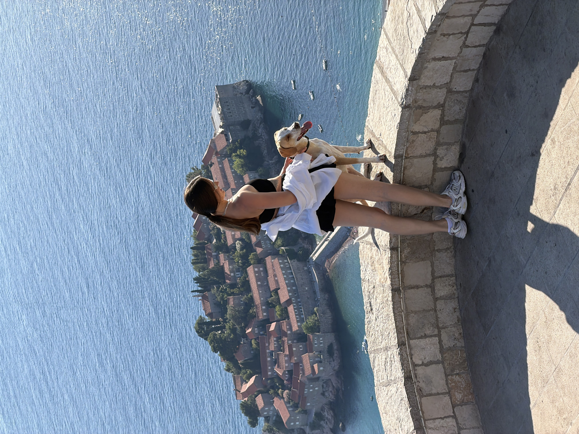 An elevated, full-body shot from behind shows a young woman standing next to a light-colored dog. The woman has long, dark brown hair and is wearing a black tank top, black shorts, and white sneakers. A white shirt is draped over her shoulders. The dog is facing right with its tongue out. They are positioned on a stone walkway with a stone wall in the foreground. In the background, the island of Sveti Stefan, Montenegro, is visible. The island is densely packed with buildings featuring red-tiled roofs and is surrounded by the blue Adriatic Sea. Several small boats are scattered on the water. The lighting suggests it is a sunny day.