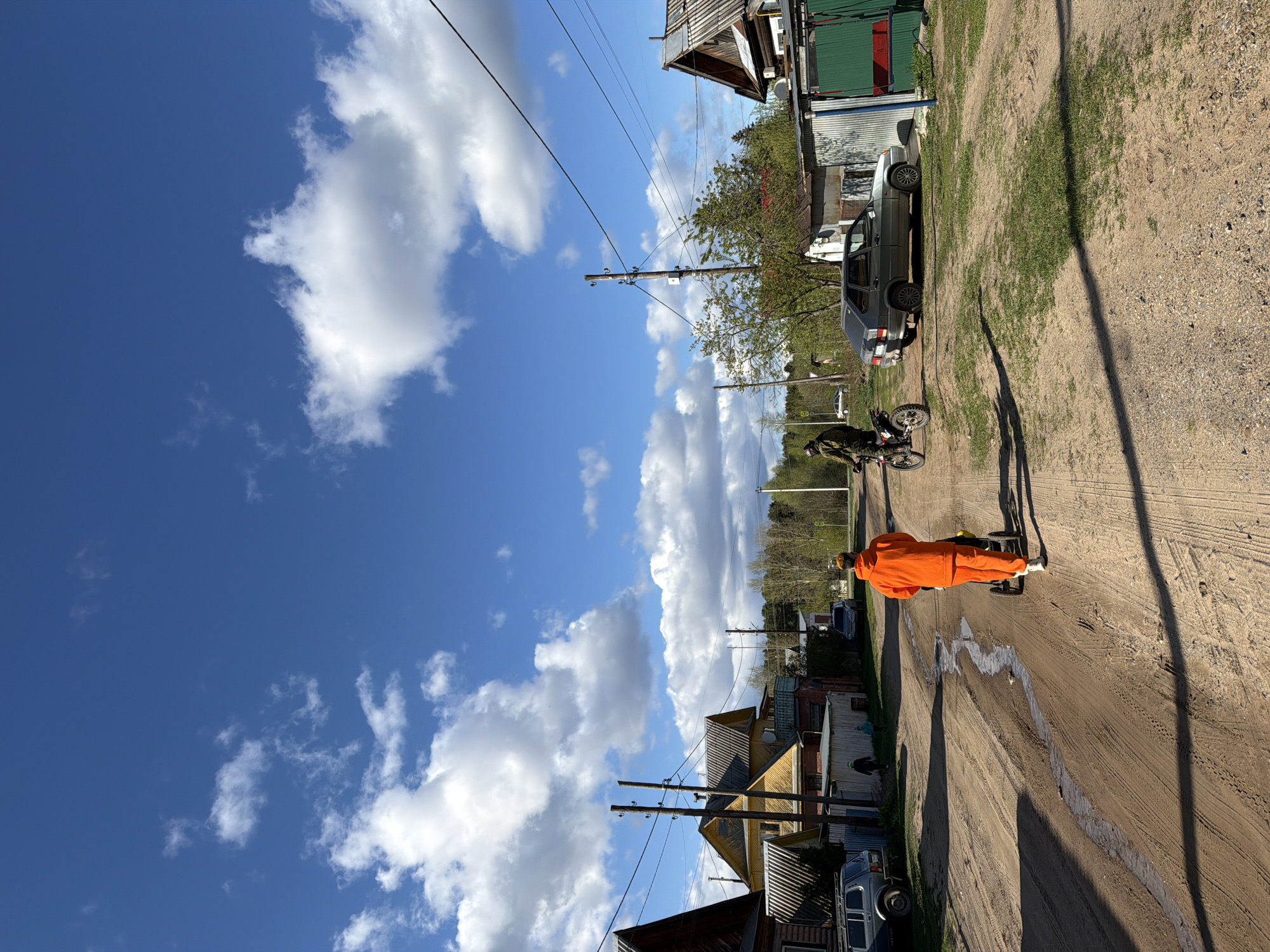 The image depicts a dirt road in Chaikovsky, Russia. A person dressed in an orange jumpsuit stands on the road, facing away from the viewer. To the left of the road are houses and parked cars. Power lines and telephone poles run alongside the road. The sky is blue with white clouds. A motorcycle is parked near a car further up the road. The road surface shows signs of recent water runoff.