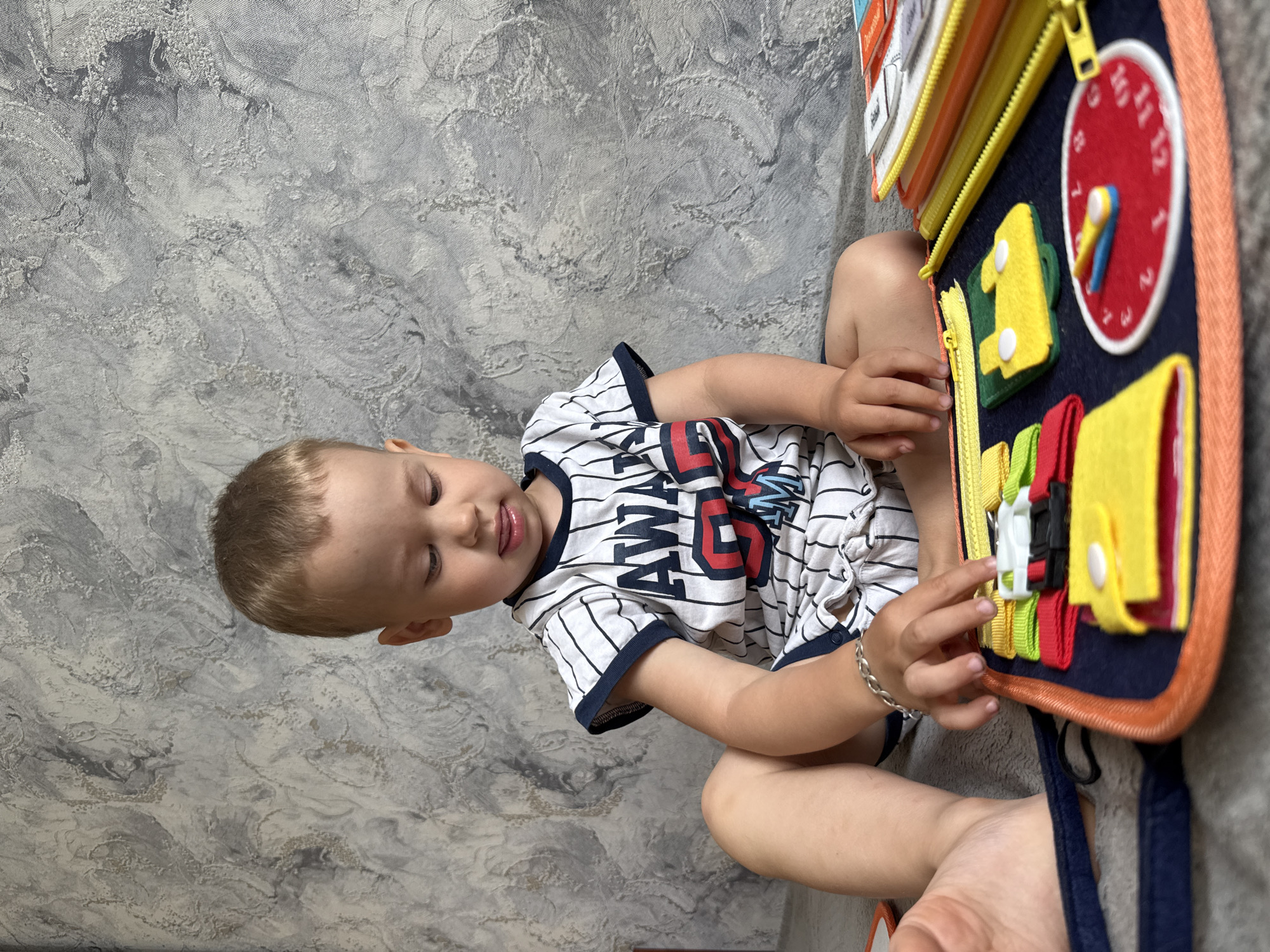 A young child sits on a gray textured surface, possibly carpet or upholstery. The child is wearing a white t-shirt with black stripes and red and blue lettering. The child is engaging with a busy board. The busy board is multi-colored and features activities such as zippers, buckles, and a clock. The background appears to be a textured wall. The location is Rusanovo, Russia.