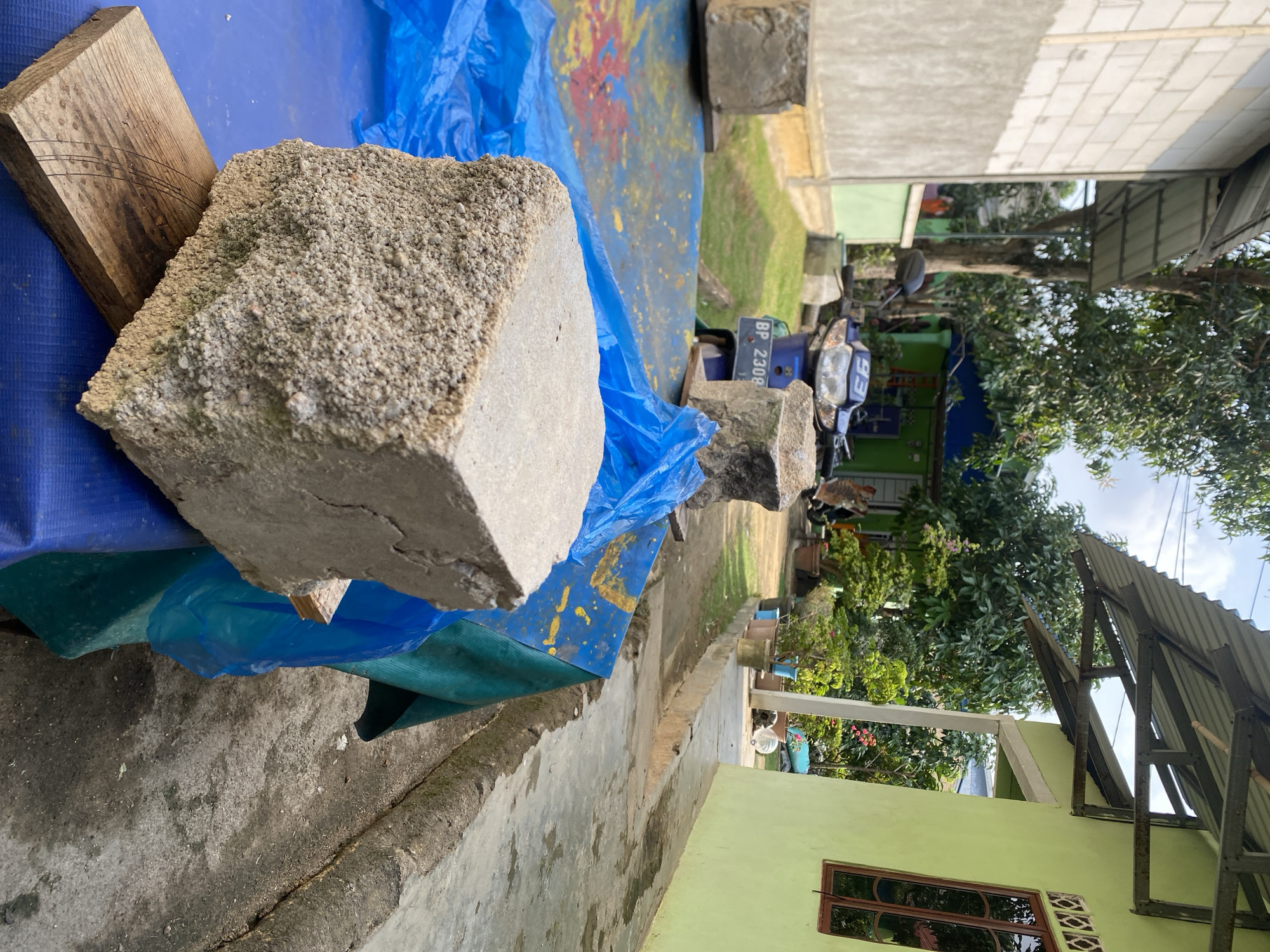 This is a photograph taken outdoors in Batam, Indonesia. In the foreground, there are two large concrete blocks, one resting on a wooden plank. Both blocks are situated on a blue tarp, which is partially covering a green tarp. In the mid-ground, a blue motorcycle with license plate BP 2308 is visible. Behind the motorcycle, there is a green building with a blue roof. In the background, there are trees and two houses, one white and one light green. The overall scene suggests a residential or construction area.