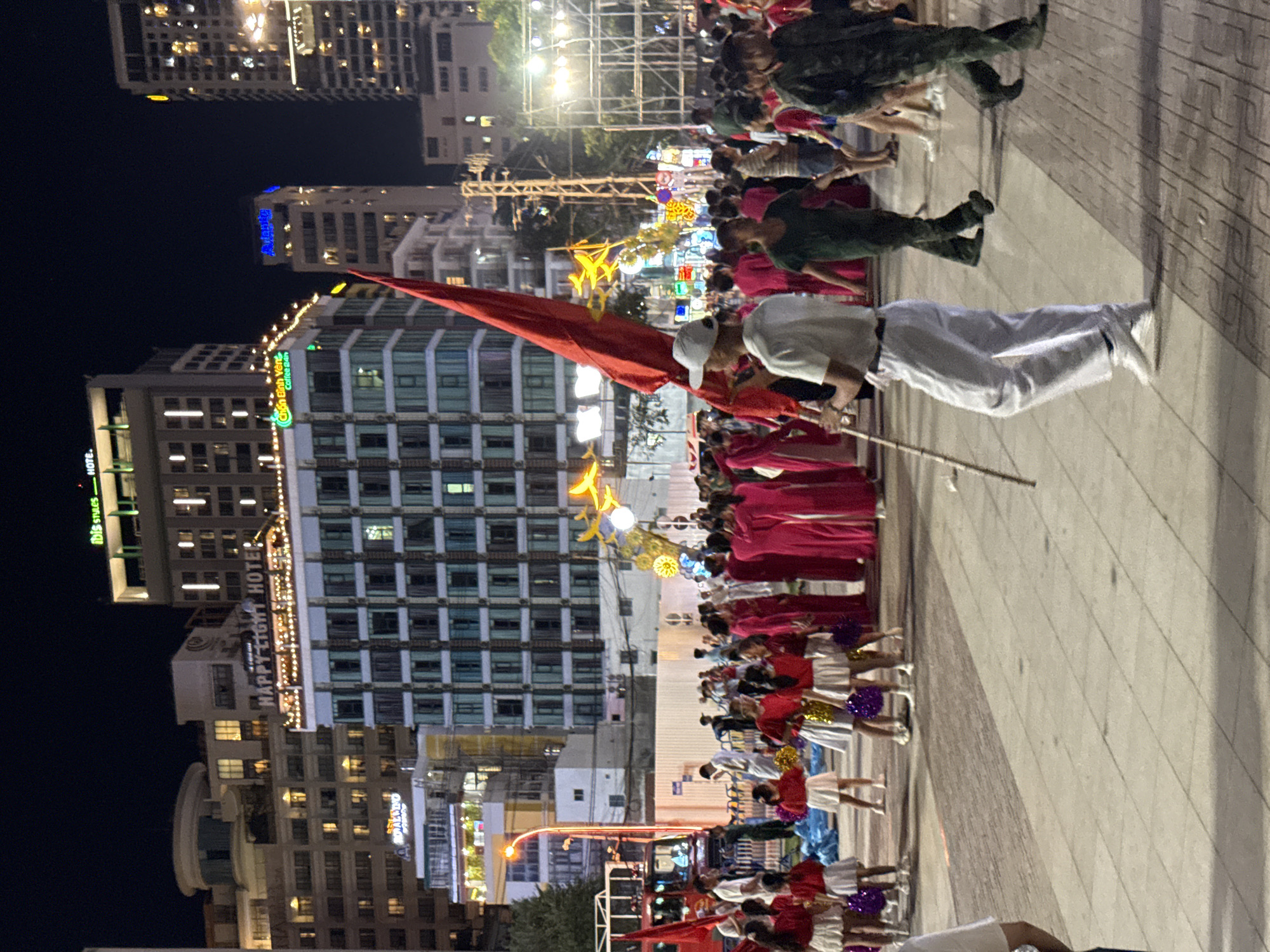 The image depicts a nighttime scene in Nha Trang, Vietnam, with a group of people participating in what appears to be a parade or performance. Several buildings are visible in the background, including the "HAPPY LIGHT HOTEL" with illuminated signage and a building displaying "ibis styles". In the foreground, a person in white attire is holding a long pole with a large red flag. They are walking on a paved walkway, casting a shadow. To the left, a group of individuals, primarily young females in red outfits and white skirts, are present, some holding pom-poms. To the right, two individuals are walking away from the camera, one in a green military-style uniform and the other in dark clothing. The environment is illuminated by streetlights and building lights, creating a festive atmosphere. Decorative lights, including star-shaped and spherical elements, are also visible, suggesting a special event or celebration.