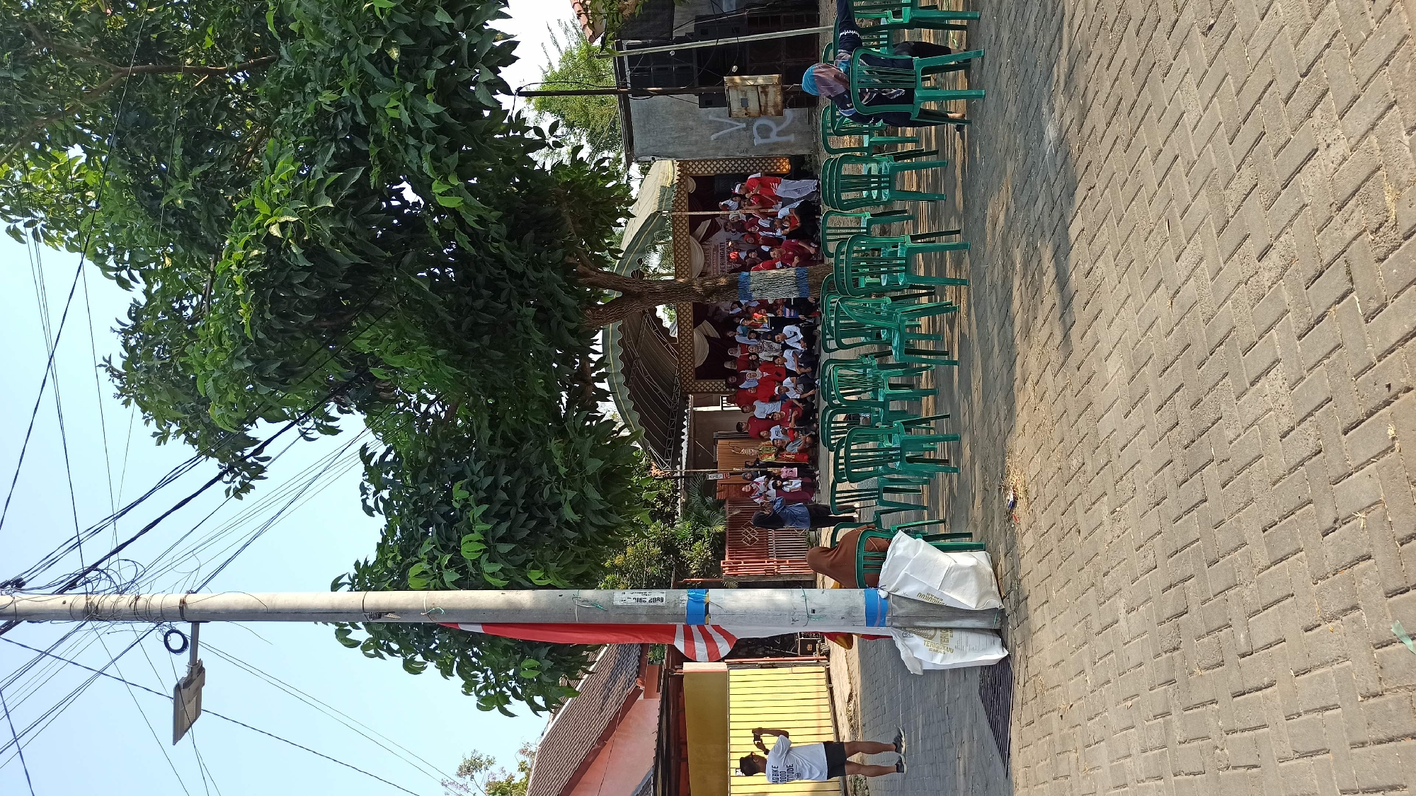 The image depicts an outdoor gathering in what appears to be a residential area. In the foreground, a man in a white t-shirt and black shorts is taking a photograph with a smartphone. He is standing next to a utility pole with a red and white flag attached to it. Rows of empty green plastic chairs are arranged on a brick-paved ground, facing a stage area. Behind the chairs, a group of people, mostly dressed in red and white, are seated or standing on and around a raised stage. A large tree with dense foliage shades part of the scene. In the background, houses with red roofs and a tent structure covering the stage are visible. A large speaker system is positioned to the right of the stage. The environment suggests a community event or celebration. The location is identified as the city of Galak, Indonesia.