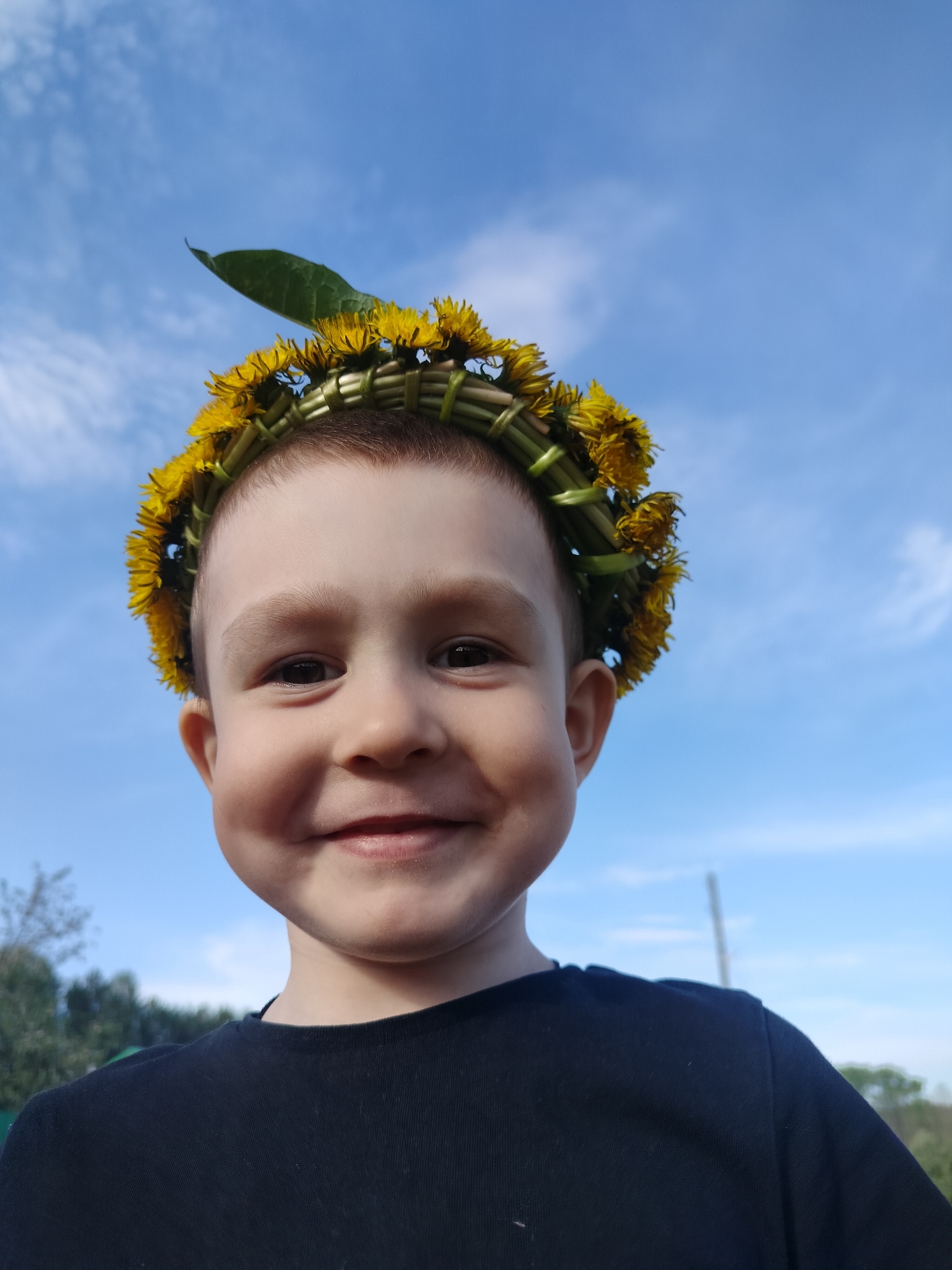 A young boy smiles at the camera, wearing a crown of yellow dandelions. The background is a blue sky with wispy white clouds. There is some green vegetation visible at the bottom edges of the frame. The boy is wearing a dark-colored shirt. The scene appears to be outdoors on a sunny day. The location is not discernible as Bormist, Russia from the provided image.
