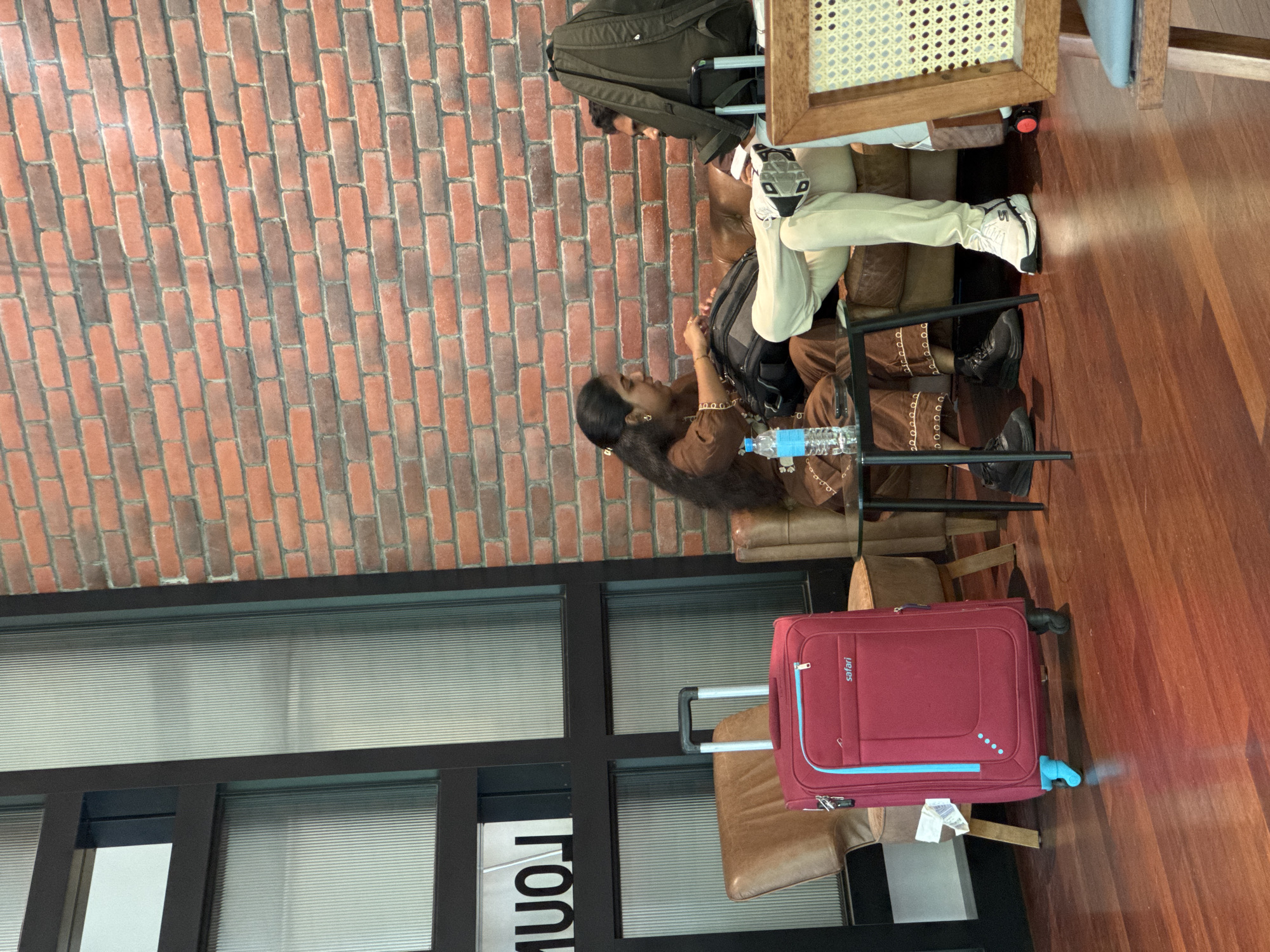 The image shows a waiting area with a brick wall in the background and a wooden floor. There are three people visible. In the foreground, a woman with long dark hair is wearing a brown outfit and is sitting on a chair, looking to her left. Next to her, on the floor, is a red suitcase with blue accents and the brand name "safari" visible. Behind the woman, a man is seated, wearing beige pants and white sneakers, with a backpack resting on his lap. Another person is partially visible in the background, also seated. The setting appears to be an indoor waiting area, possibly an airport lounge or a cafe, given the presence of luggage and seating arrangements. The lighting suggests it is daytime, and there are no visible weather cues. The overall atmosphere appears calm and relaxed, with the individuals seemingly waiting.