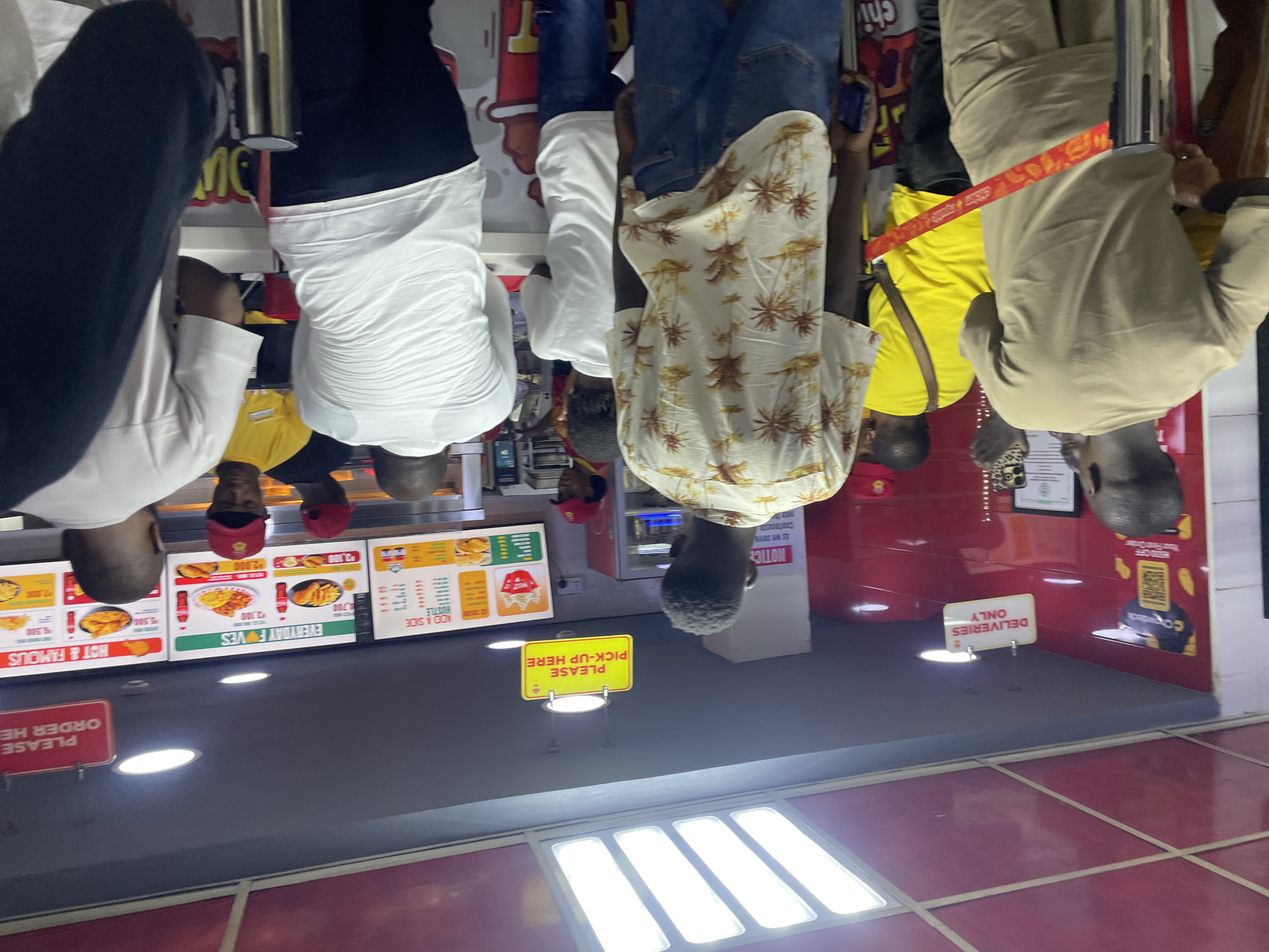 This image captures an indoor scene within a fast-food restaurant in Abuja, Nigeria. The original photograph is rotated 90 degrees counter-clockwise; when viewed upright, people are seen standing on a grey floor.
Several individuals, mostly men, are present, appearing to be queuing or waiting for service. On the left, a man in a white long-sleeve shirt and dark pants stands near a counter. In the center, a man wears a white short-sleeve shirt with a brown palm tree pattern and blue jeans. To the right, another man in a light brown long-sleeve shirt and light brown pants stands behind a silver metal barrier. Other patrons or staff are partially visible, including individuals in yellow shirts and one wearing a red cap.
The setting features prominent red walls with a shiny finish, and bright recessed lighting illuminates the space from both the ceiling and what appears to be the floor or a lower counter. Various signs are displayed: a red sign on the far left reads "PLEASE ORDER HERE," while a yellow sign in the middle states "PLEASE PICK-UP HERE." A white sign on the right indicates "DELIVERIES ONLY." Menu boards behind the customers advertise items like "HOT & FAMOUS" and "EVERYDAY FOVES" with prices in the thousands, such as "2,000," "2,100," "3,700," and "4,500." Other visible text includes "HUSTLE," "ADD A SIDE," "chi" (a brand name), and a "NOTICE" on the wall. The scene suggests a busy quick-service restaurant environment where customers are processing orders.