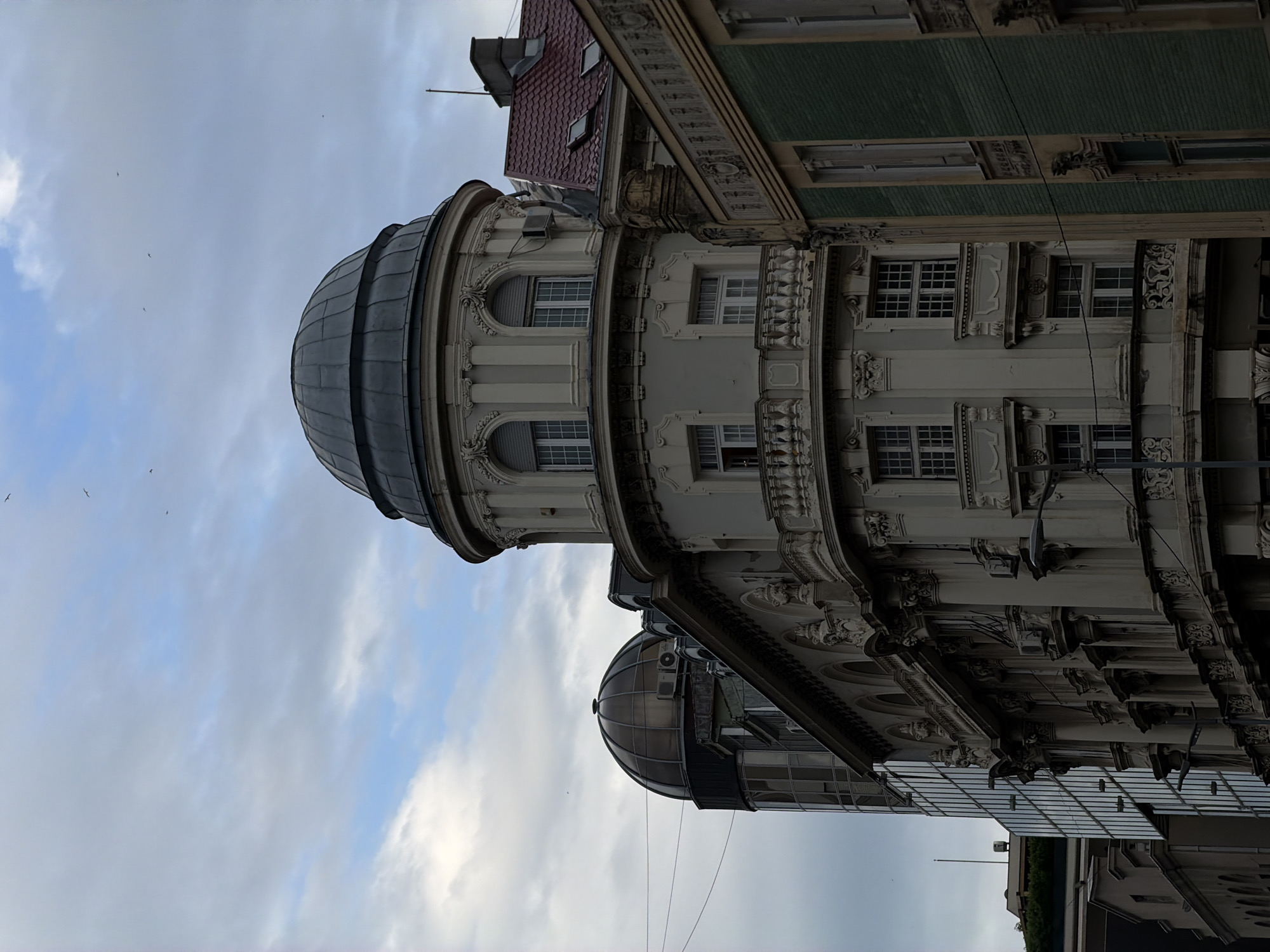 This is a low-angle, full-shot image of a street in Belgrade, Serbia, featuring a cluster of diverse architectural styles. The dominant structure in the foreground is a light gray building with ornate classical detailing, featuring a prominent rounded section topped by a dark dome. This building has multiple windows, some with balconies supported by decorative brackets. To its left, another building with a glass facade rises, and further to the left, a dark, rounded dome structure is partially visible. To the right of the main building, a structure with a red tiled roof and dormer windows is visible, adjacent to a building with a distinctive green tiled facade. The sky above is overcast with scattered clouds, and several birds are in flight. The scene depicts an urban environment with a mix of old and new architectural elements.