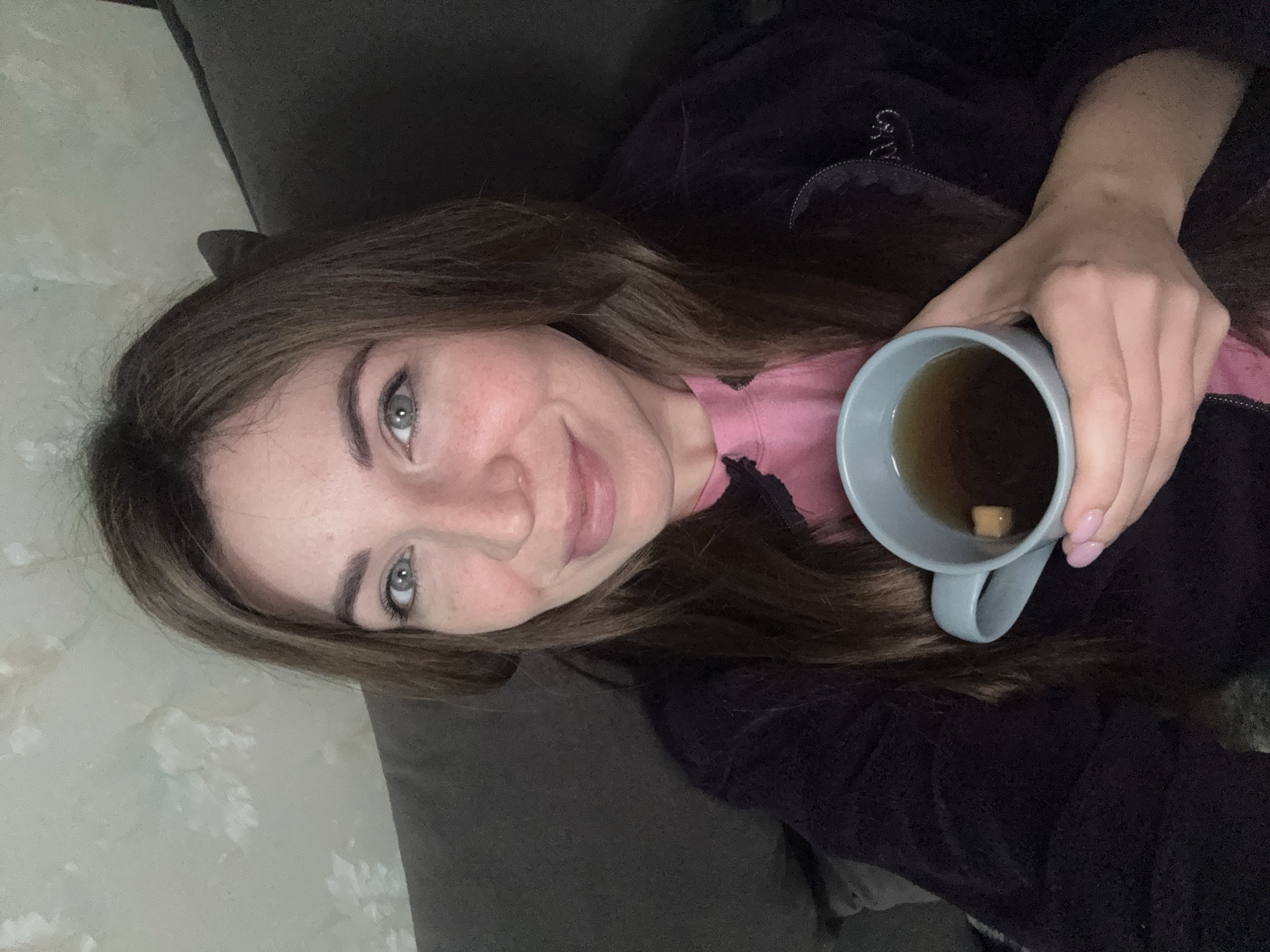 A woman is holding a mug containing a brownish liquid. She is wearing a pink top under a dark-colored jacket. The setting appears to be indoors, with a couch visible in the background. There is no indication of the location in Kryvyi Rih, Ukraine, from the image.