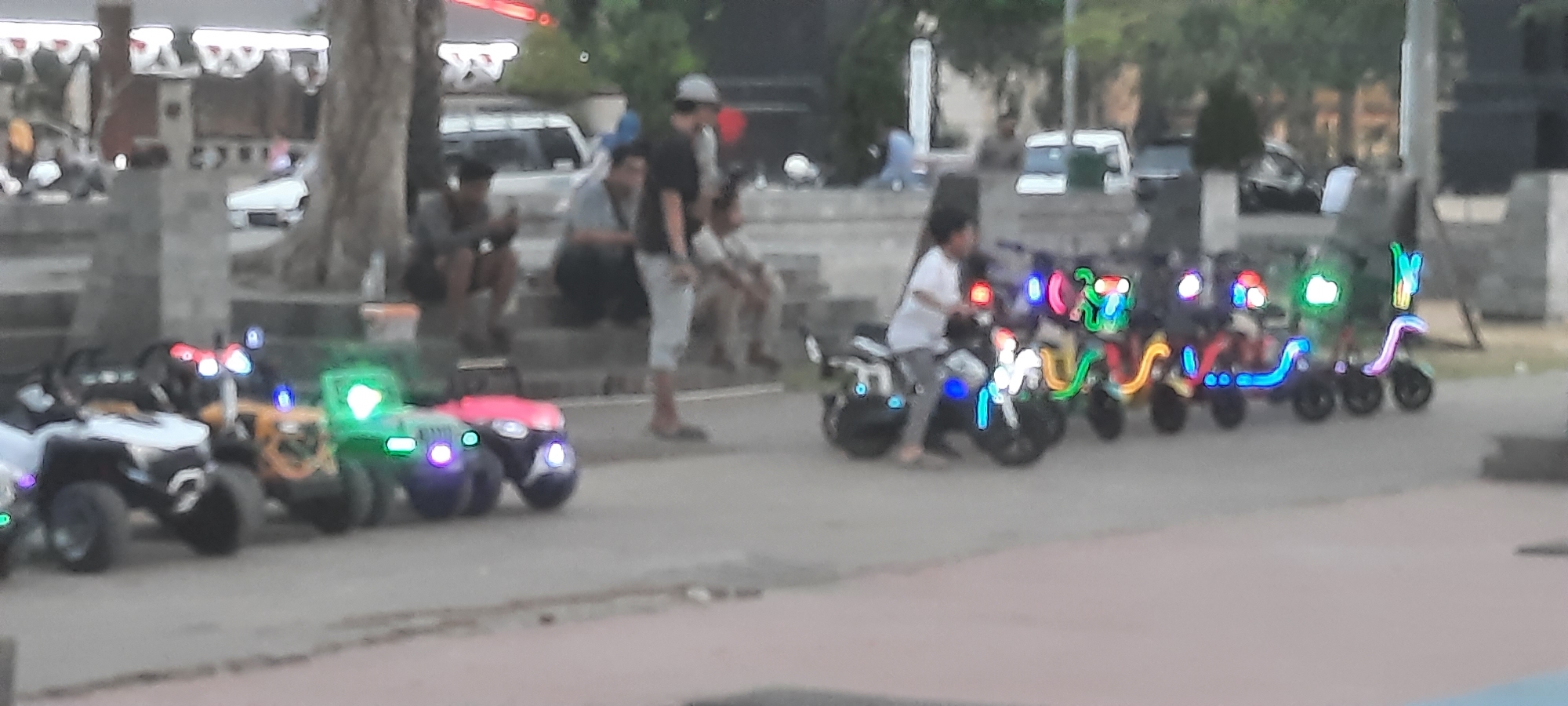 A low-angle, medium shot captures a line of brightly lit electric toy vehicles, resembling scooters and mini-jeeps, on a paved walkway. The toys are decorated with an array of colorful LED lights in vibrant blues, greens, pinks, and yellows, creating a festive atmosphere. A child, wearing a white shirt and gray pants, is seated on one of the electric scooters, appearing to operate it. Several other toy vehicles, some with additional lights and designs, are parked in a row behind the child's scooter. In the background, a group of individuals is seated on concrete steps, some looking towards the toy vehicles. The setting appears to be an outdoor public space, possibly a park or a recreational area, with trees and structures visible in the distance. The overall impression is one of a lively, family-friendly environment where children can enjoy playing with illuminated toys. The image is blurry due to a shallow depth of field and low resolution, suggesting it was taken with a mobile phone camera under low-light conditions. The location is identified as the city of Kanigoro, Indonesia, specifically near the city's administrative center or a local park where such attractions are common.