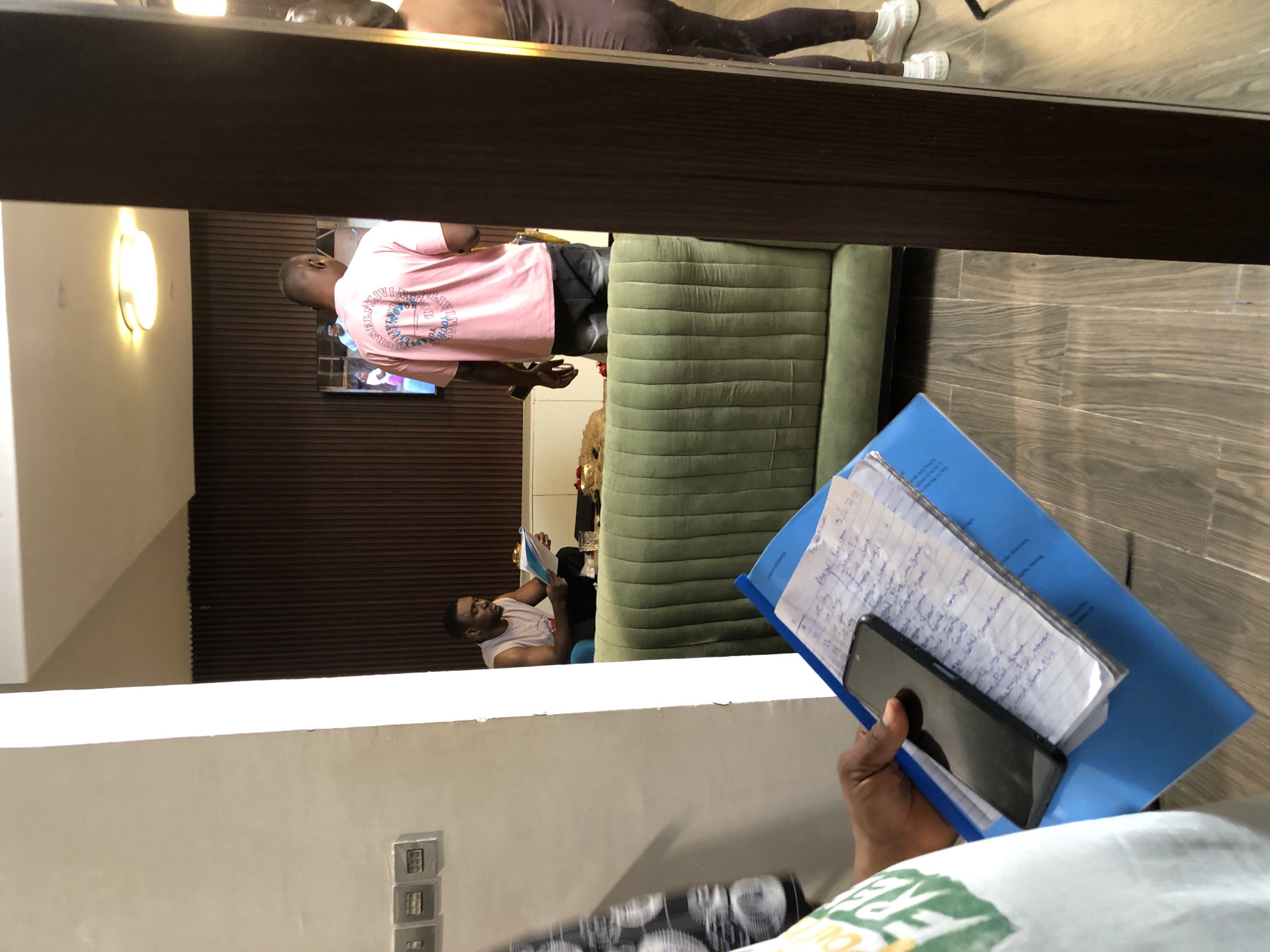 The image captures an indoor scene from a slightly elevated, first-person perspective, looking down into a lounge or waiting area. In the immediate foreground, a person holds a blue folder containing handwritten papers and a black smartphone. The hand holding these items is dark-skinned, and part of a light-colored t-shirt with green and yellow text is visible.
In the mid-ground, below a dark horizontal beam, a man stands facing away from the camera, wearing a pink t-shirt with a circular graphic and text on the back, and dark shorts. The text on his shirt includes phrases like "FEEL ALIVE" and "FIND JOY IN THE JOURNEY." He has short hair and appears to be observing something ahead of him, possibly a partially visible television screen on a dark, wood-slatted wall. To the right of him, a long, green, tufted sofa stretches across the frame.
Further into the room, to the left of the sofa, another man is seated on a blue chair, engrossed in reading a book or papers he holds. He wears a light-colored tank top and dark trousers. Behind him, some decorative items, including a gold-colored piece, are visible on a surface. The room features light-colored walls, one of which has an oval-shaped light fixture emitting warm light, and another with three silver rectangular wall outlets/switches. The floor is tiled with a wood-grain pattern.
A notable detail is a reflection visible above the standing man, showing the lower body of a person wearing dark leggings and white sneakers, as if looking into a mirror or highly polished surface. The handwritten papers in the blue folder show words like "Myself," "plan," "daily," "Monday," and a date such as "9-2-20." The overall lighting is bright, suggesting daytime or a well-lit interior.