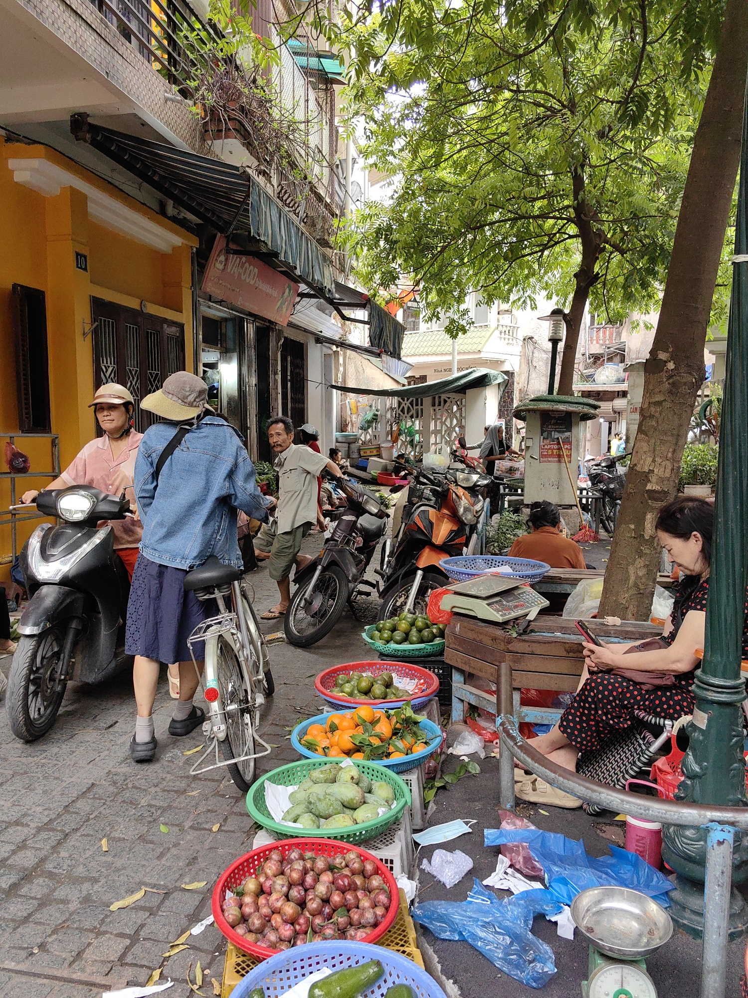 This daytime image depicts a lively street market scene in Hanoi, Vietnam. The narrow street is filled with people, vehicles, and fresh produce, creating a vibrant atmosphere.
In the foreground and middle ground, several individuals are engaged in daily activities. On the left, a woman in a pink shirt and a light-colored cap sits on a black scooter. Beside her, another woman wearing a denim jacket, a dark blue skirt, and a wide-brimmed hat stands next to a bicycle. Further right, a man in a light shirt and shorts stands near a parked motorbike, looking towards the left. On the far right, a woman in a black and red polka-dot dress is seated on a low wooden bench, looking at her mobile phone. Another person is partially visible sitting further back, obscured by a tree trunk.
The ground is arranged with colorful plastic baskets containing various fresh fruits, including red plums, green mangoes, bright oranges, and small green limes or similar citrus fruits. An electronic scale rests on a wooden crate next to some of the baskets, indicating sales are taking place. A traditional metal scale is also visible on the ground. Several blue plastic bags, some discarded litter, and fallen leaves are scattered on the paved street.
The street is lined with buildings, including a prominent yellow structure with the number "10" clearly visible on its facade. A red sign above a storefront displays "VAI-FOOD". Retractable awnings extend from several buildings, providing shade. A large tree with dense green foliage stands tall on the right, its trunk forming a significant part of the frame. A utility box in the mid-ground features text such as "LAPTOP PC ĐIỆN THOẠI" (Laptop PC Phone) and "ĐÔNG NAM Á" (Southeast Asia). The overall scene captures the bustling street life and commerce typical of Southeast Asian cities.