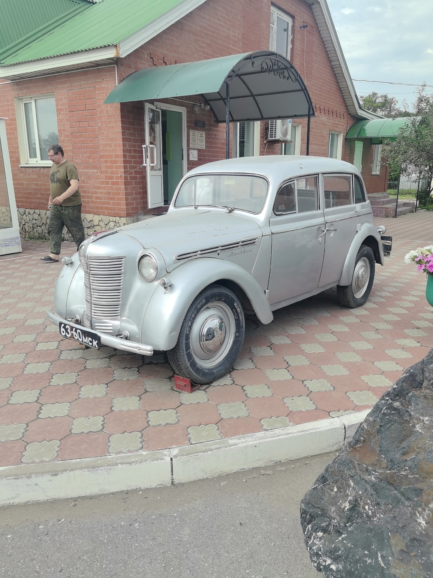 A silver vintage car is parked on a patterned brick walkway in front of a red brick building with a green roof. A man wearing a green t-shirt and camouflage pants walks away from the car. The car has a license plate that reads "63-60MCK". The building has a green awning over a doorway. The scene appears to be outdoors, possibly in a small town or rural area in Russia.