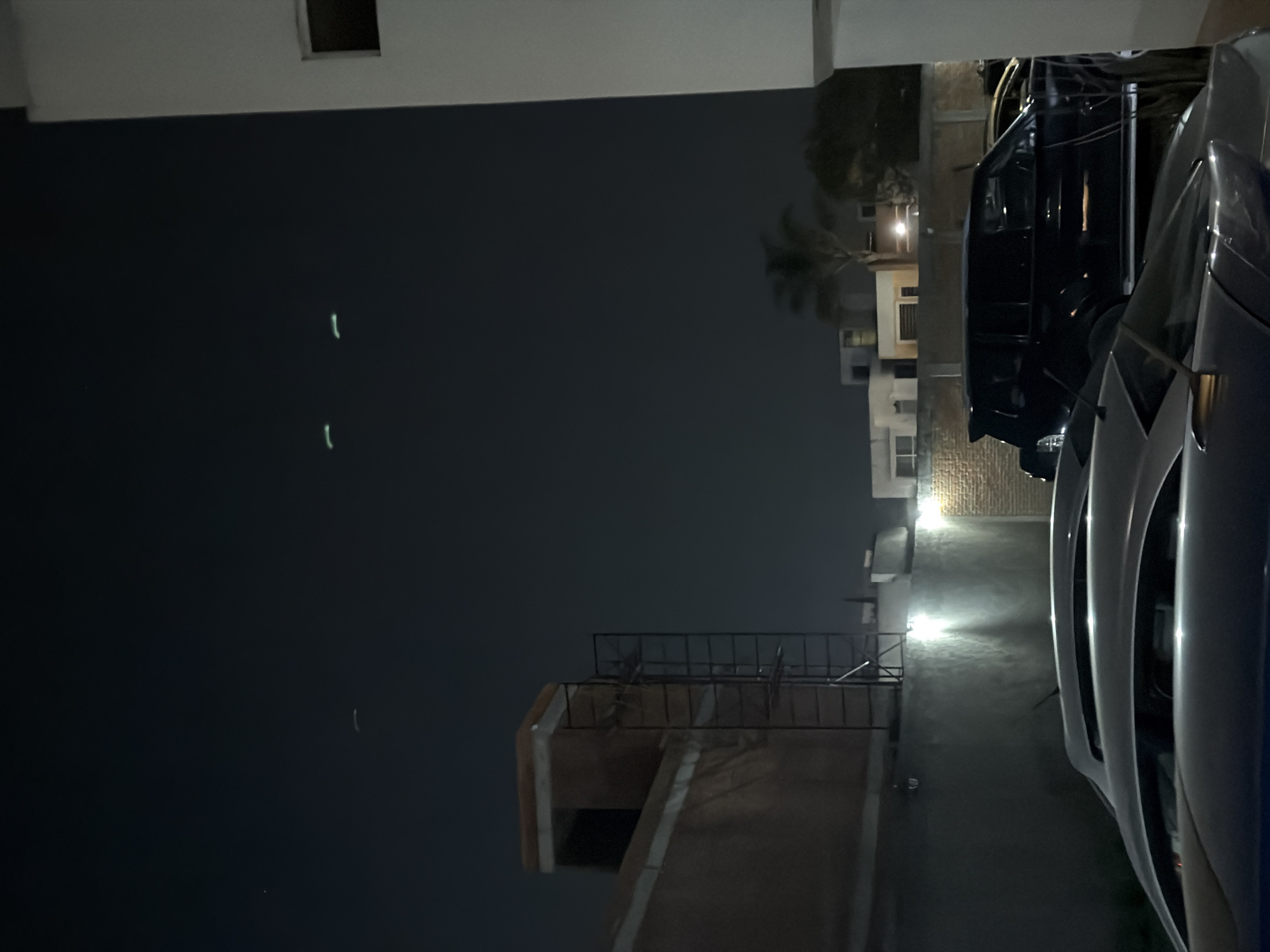 This is a night photograph taken outdoors, likely in an urban or suburban setting. The scene is illuminated by artificial light sources.
In the right foreground, parts of two cars are visible. The car closest to the viewer is light-colored, possibly silver or white, showing its right front fender and side profile. Behind it, a darker car, likely black or dark blue, is also parked. Both cars appear to be facing towards the left of the frame.
Towards the midground on the left, a multi-story building with a flat roof and a railing is visible. The architecture suggests a residential or commercial structure. A bright light source on or near this building illuminates the ground and part of a nearby wall. Another building with windows and a palm tree are visible further back on the left side of the image, also illuminated by scattered lights.
The sky, occupying a significant portion of the upper left, is dark, indicating nighttime. Notably, there are two distinct, elongated, bright green-white light streaks suspended vertically in the upper left part of the dark sky. A smaller, fainter light point is also present below them. The overall impression is calm and static, with no human activity evident.
There is no visible text in the image. The setting, with its specific architecture and palm tree, is consistent with the provided location context of San Andrés Cholula, Mexico.