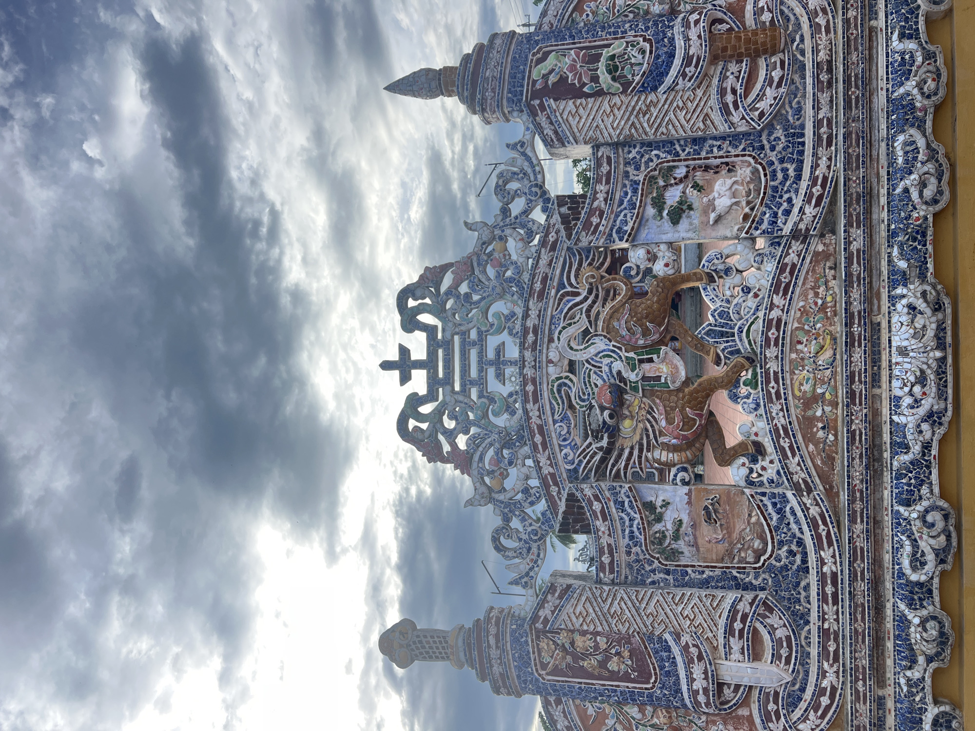 The image displays an elaborately decorated facade, likely a tomb or temple entrance, located in Hoi An, Vietnam. The structure is adorned with intricate mosaic tiles depicting various motifs. Prominently featured are two stylized, horse-like creatures in a dynamic pose, rendered in a reddish-brown hue. These figures are set against a backdrop of blue and white patterned tiles, which also form decorative borders and panels.
The facade is further embellished with other mosaic scenes. To the left of the central figures, there is a depiction of a person plowing a field with an animal, possibly a water buffalo, and further to the right, a scene of a person riding a white animal, resembling a goat or deer, in a pastoral setting. Above these scenes, ornate ceramic designs in blue, white, red, and green create a visually rich and complex pattern, including floral elements and what appears to be a stylized Chinese character or symbol at the apex. The entire structure is topped with decorative spires and finials.
The sky above is overcast, with dramatic grey clouds illuminated by a bright, diffused light, suggesting either early morning or late afternoon. The architectural style and the vibrant mosaic work are characteristic of historical Vietnamese tombs and temples, particularly those found in central Vietnam, including the imperial city of Hue and its surrounding areas, and are also found in notable structures within Hoi An. The overall impression is one of rich cultural heritage and artistic craftsmanship.