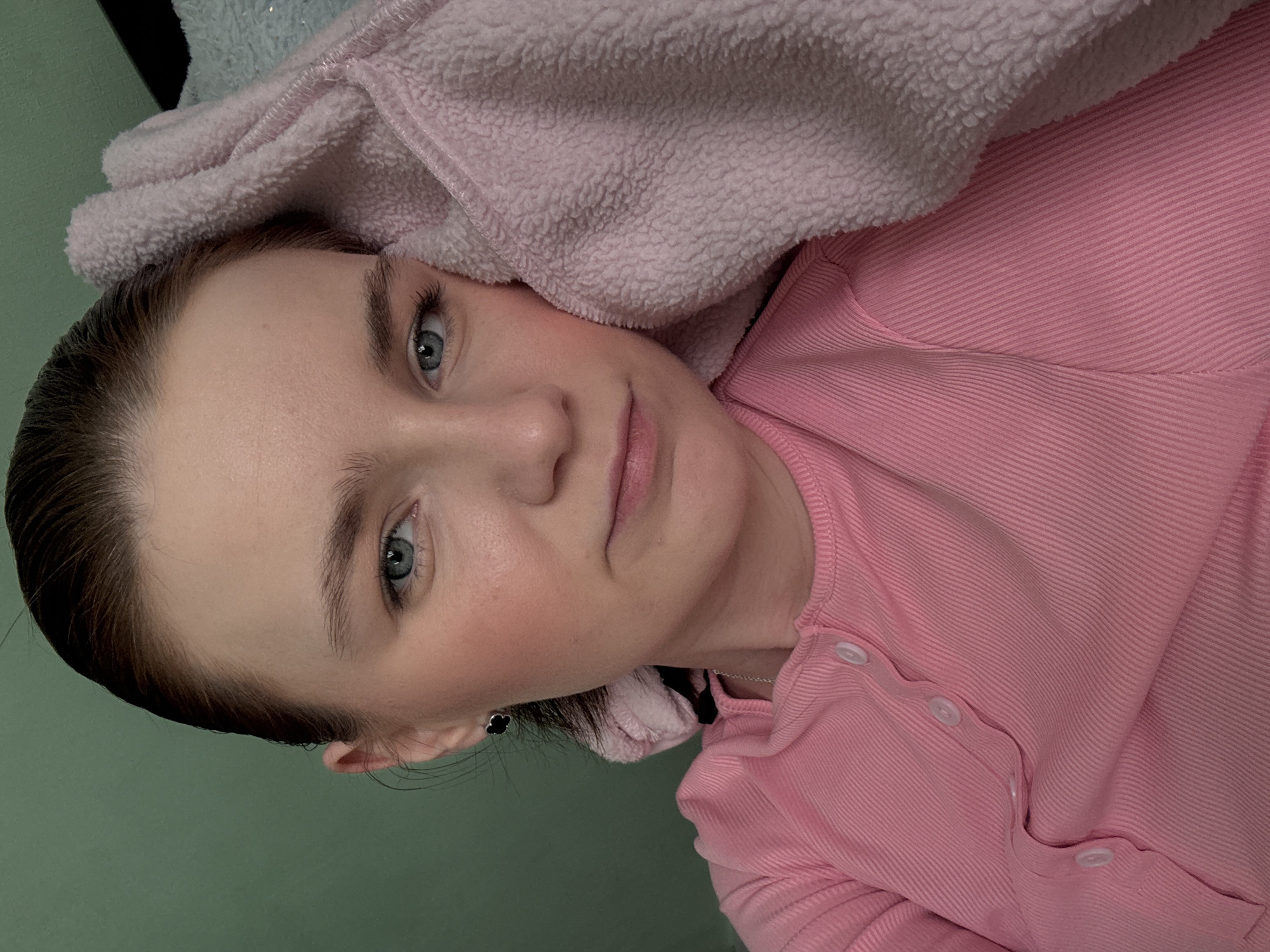 A woman with light skin and blue eyes is pictured in a selfie. She is wearing a pink, ribbed top with white buttons. A light pink, soft, fleece-like material is draped over her head. She is facing the camera with a neutral expression. Her hair is dark and slicked back. She is wearing a small, dark earring. The background is a light green wall. The location and date are unknown.