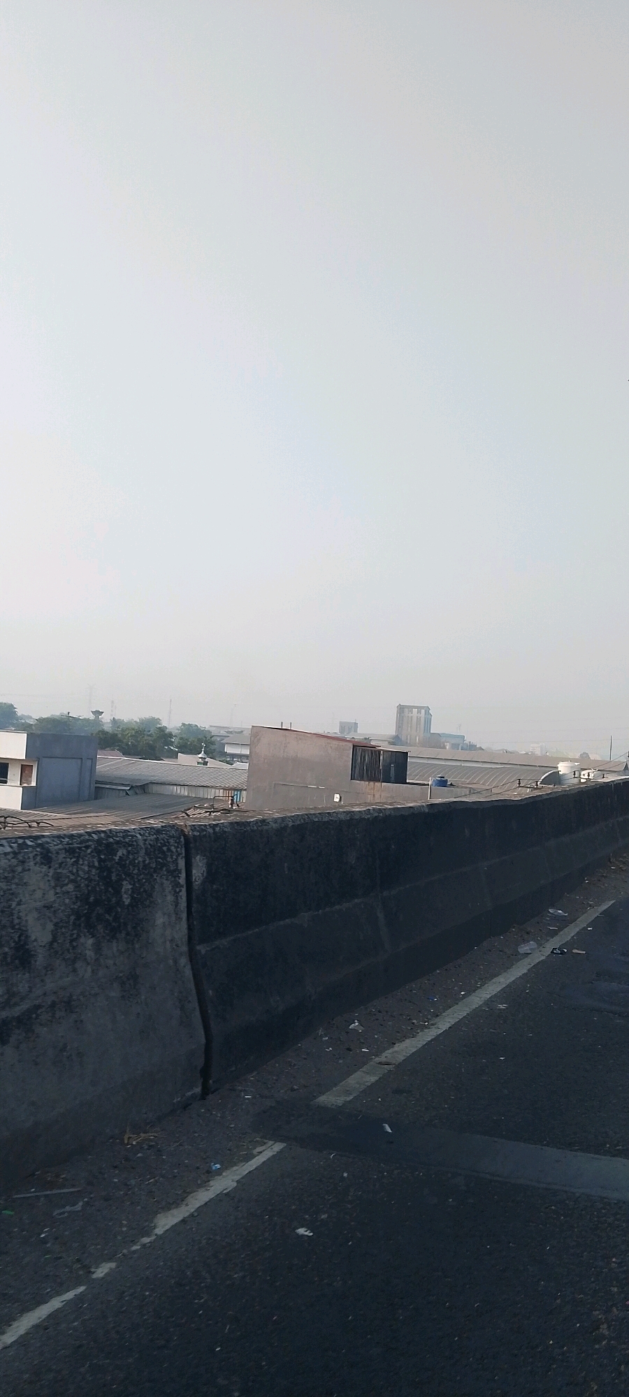 The image captures a view from a highway overpass in Bekasi, Indonesia, looking out over an industrial area. In the foreground, a concrete barrier separates the roadway from the industrial zone. The barrier is weathered and shows signs of wear and tear, with some debris scattered along its base. The roadway itself is asphalt, with white lane markings visible.
Beyond the barrier, a collection of industrial buildings dominates the landscape. These structures are characterized by their varied shapes and sizes, featuring corrugated metal roofs, concrete walls, and some with visible ventilation systems. One building has a distinctly angled, grey facade with a grated section at the top. In the mid-ground, a tall, slender water tower stands out against the hazy sky, a common feature in industrial districts. Further in the distance, more buildings are visible, including what appears to be a taller, multi-story structure, suggesting a more developed part of the city.
The sky is overcast and hazy, a common atmospheric condition in urban and industrial areas, potentially indicating smog or pollution. The overall scene conveys a sense of urban sprawl and industrial activity. There are no people visible in the image, and the focus is solely on the environment and structures. The light is diffused, suggesting it might be early morning or late afternoon.