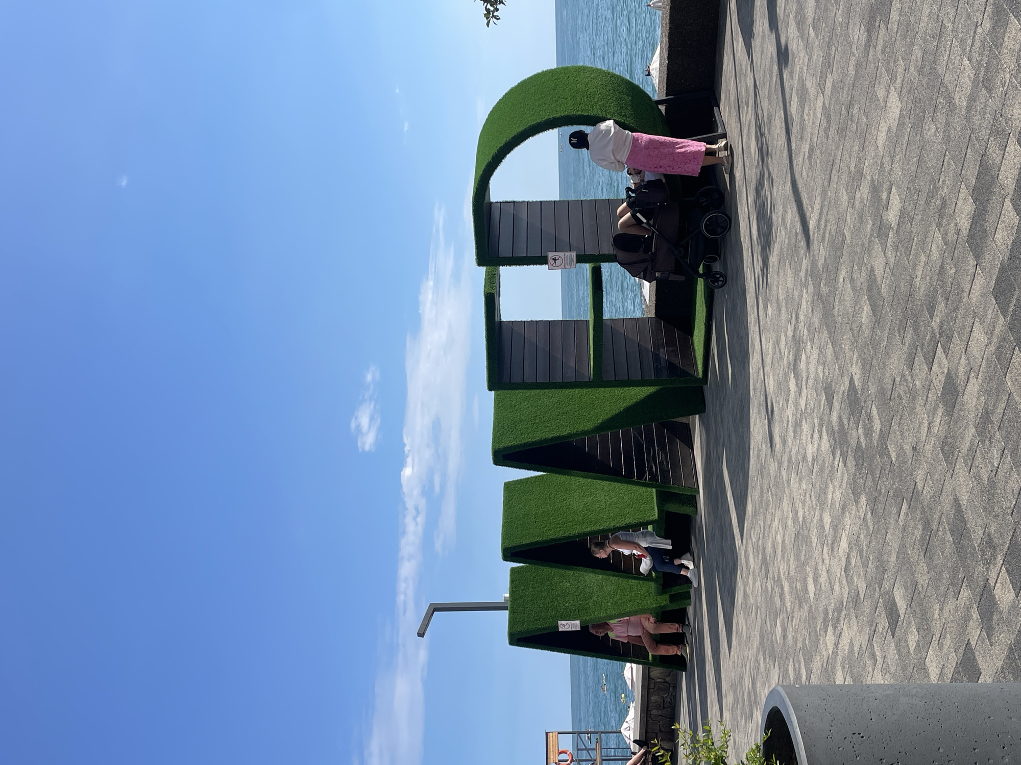 The image depicts a public space along the coastline in Sochi, Russia, featuring a large, decorative installation spelling out "MAD" in bold, green letters. The letters are constructed with a dark, wood-like material on the sides and back, and covered in artificial grass on the front and top, creating a visual contrast with the bright blue sky and the sea.
In the "M" section of the installation, two individuals are seated on benches: a man in a pink t-shirt and light-colored pants, and a woman in a white tank top and jeans. They appear to be resting and observing the surroundings.
The "A" section is empty, though it features a sign with a no-smoking symbol.
The "D" section contains a woman in a white top and a long pink skirt, standing and tending to a stroller. Another person's legs are visible inside the stroller. This section also has a sign with a no-smoking symbol.
The ground in front of the installation is paved with interlocking grey bricks, creating a geometric pattern. In the foreground, to the left, a portion of a large, cylindrical grey planter is visible, containing green foliage.
The background reveals the Black Sea, with a clear blue sky above. Several small boats or rafts are scattered on the water, and a lifeguard tower with a red ring is partially visible on the left side of the image. The overall scene suggests a leisurely day at a seaside resort.