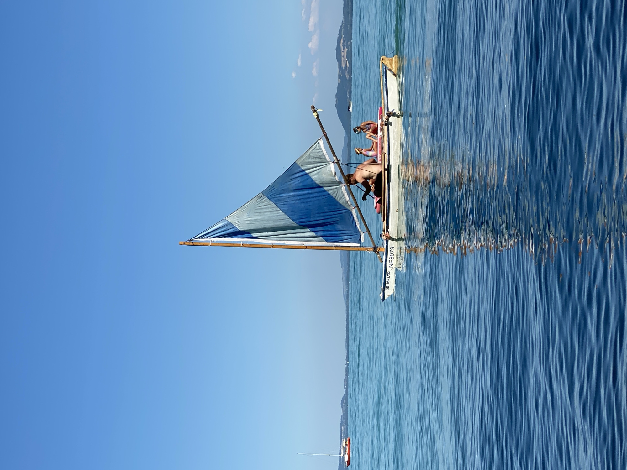 A small sailboat with a blue and white sail is sailing on a clear blue lake under a cloudless sky. Three people are aboard the sailboat: an adult male is at the helm, and two children are seated on inflatable mats. The sailboat has the registration "NE8079" on its hull. In the background, another sailboat with a red hull is visible on the water, and hills form the distant horizon. The water is calm and reflects the sky and the sailboat. The scene appears to be in the vicinity of Cortaillod, Switzerland, as indicated by the boat registration.