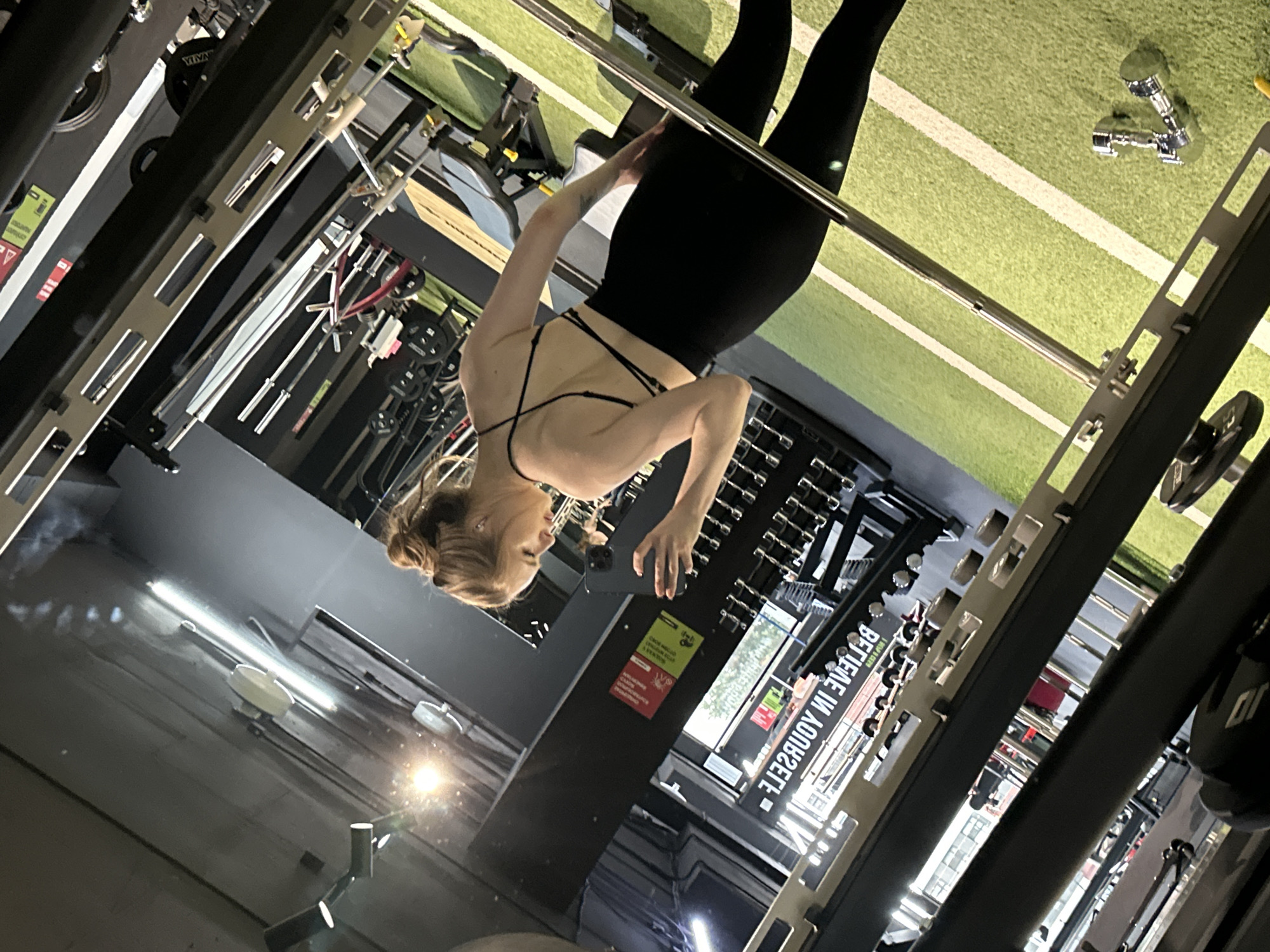 An indoor scene depicts a female in a gym or fitness center, capturing a mirror selfie. The subject, an adult female with light brown hair tied in a bun, wears a black sports bra with a multi-strap back design and black leggings. She holds a dark-colored smartphone with her left hand, positioned to reflect her image and the gym environment in a large mirror. Her gaze is directed towards the phone/mirror, and she exhibits a slight smile.
The gym floor features green artificial turf with white demarcation lines. Various pieces of exercise equipment are visible: a large, multi-functional strength training machine (possibly a Smith machine or power rack) with silver and black components frames the foreground and midground. In the reflection, a dumbbell rack with multiple sets of dumbbells, a black and grey weight bench, and additional barbells and weight plates are discernible. Two small, chrome-plated dumbbells rest on the green turf to the right of the frame.
The walls and ceiling are dark-colored. Wall-mounted and ceiling-mounted light fixtures provide illumination, with some bright light sources causing reflections. Reversed text, "BELIEVE IN YOURSELF," is clearly visible in the mirror reflection, confirming the reflective surface. Below this text, unreadable red and yellow informational signs are also reflected. The overall setting is an equipped fitness facility in the city of Unknown, Unknown.