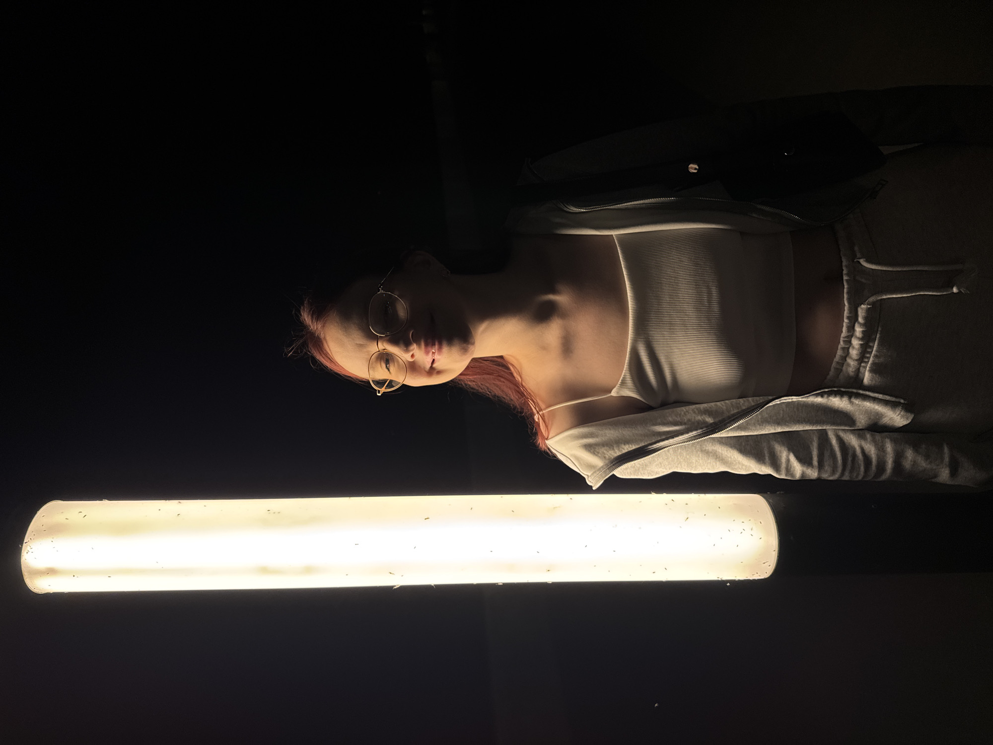 A woman with red hair and glasses is lying down in a dark room, illuminated by a fluorescent tube light in the foreground. She wears a white tank top and a grey zip-up hoodie. The background is predominantly dark. Small particles are visible on the surface of the light tube. The location is unknown and there is no evidence in the image to suggest it is Nizhny Novgorod, Russia.
