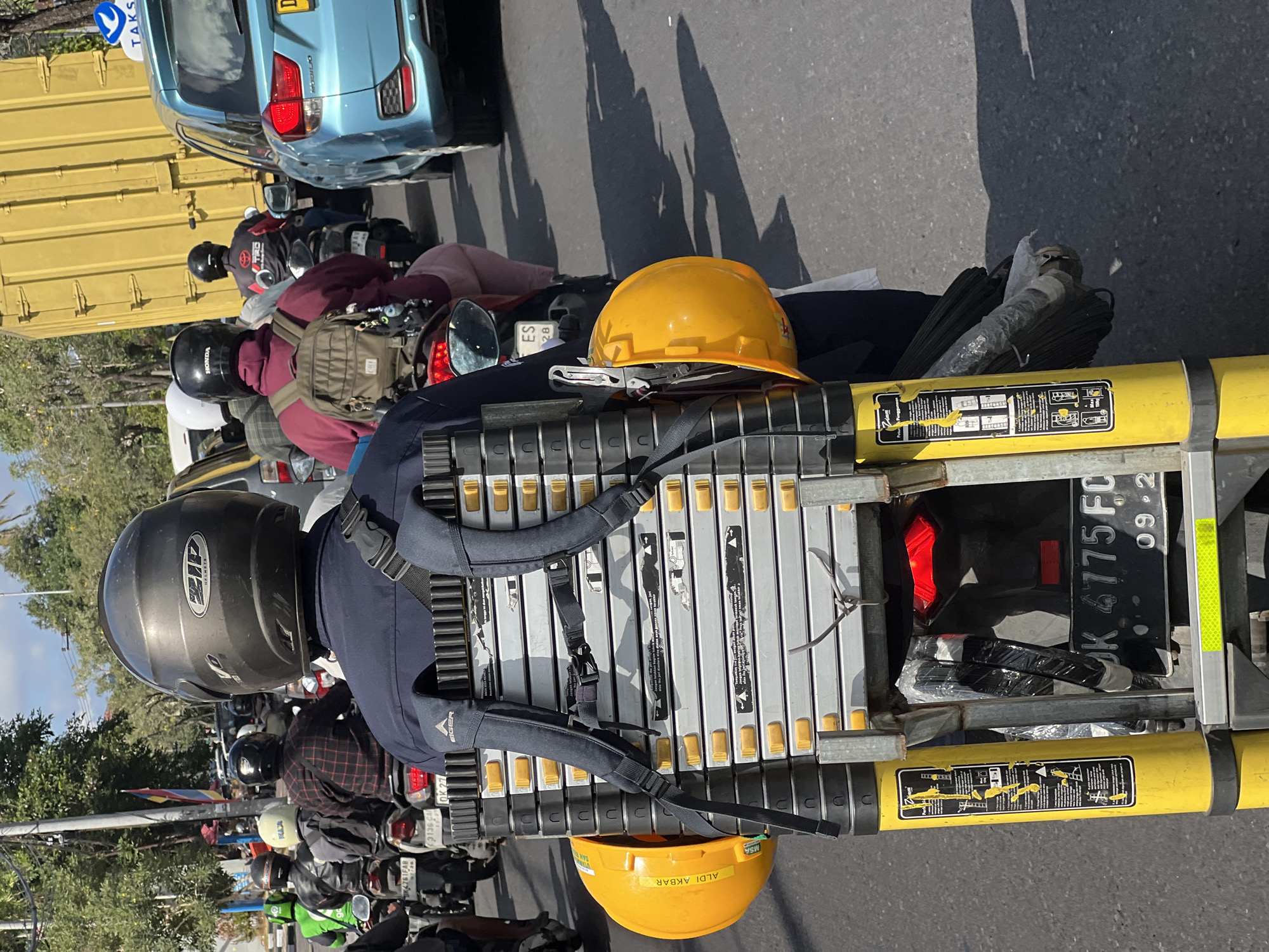 The image captures a street scene in Kuta, Indonesia, showing a man riding a motorcycle. He is wearing a dark blue uniform and a black helmet. Strapped to the back of his motorcycle is a large, multi-sectioned ladder, secured by straps from a backpack. Two yellow hard hats are attached to the ladder's frame. In front of him, other motorcyclists are also visible, some wearing helmets and backpacks. To the right, a blue car and a large yellow truck are present in traffic. The road is paved, and trees line the background. The overall impression is of a busy urban environment with typical traffic flow.