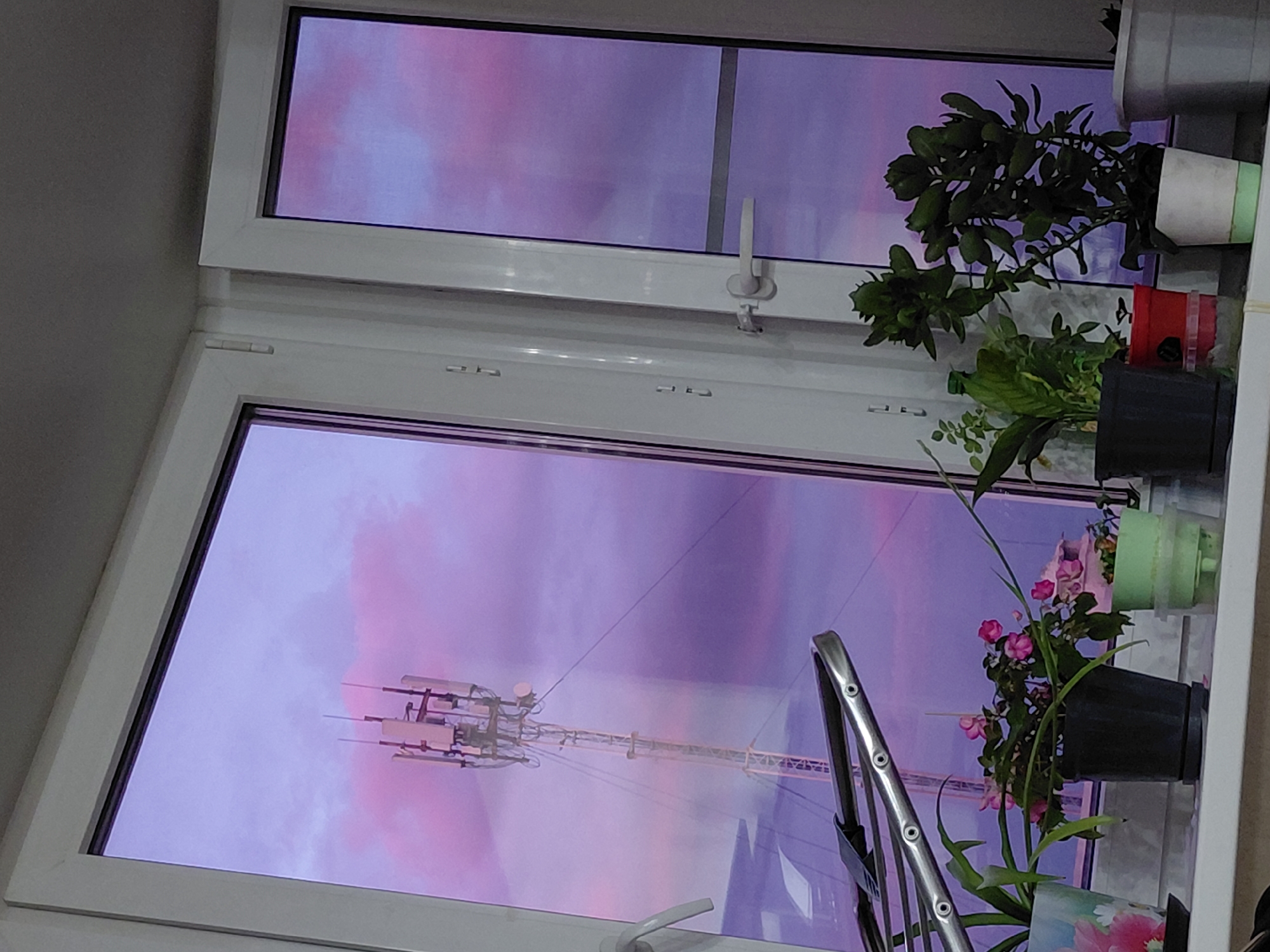 The image captures an indoor perspective looking out through a white-framed window, revealing a vibrant and colorful sky. The sky is dominated by striking shades of purple and pink, strongly suggesting either a dramatic sunrise or sunset. A tall cell tower or communication mast, equipped with multiple antennas and dishes, is prominently visible through the lower window pane, silhouetted against the intense sky colors.
The window itself is a modern, white PVC-framed unit, appearing to consist of at least two sections. A white handle is discernible on the upper window frame. To the right, on an interior windowsill or ledge, several potted plants are arranged. These include a lush green plant in a white pot with a light green rim, another green leafy plant in a black pot, and a flowering plant with small pink blooms and long green leaves, also in a black pot. A small red object and a light green inverted container (possibly for watering) are also visible among the plants. Part of a silver, metallic object, likely a household item, is visible in the lower right corner.
The scene evokes a sense of quiet observation from indoors, highlighting the contrasting elements of the natural sky, the industrial cell tower, and domestic plant life. No human figures or discernible text are present in the image. The window design and the presence of the communication tower align with the context of Nadym, Russia, likely reflecting an urban or suburban setting.