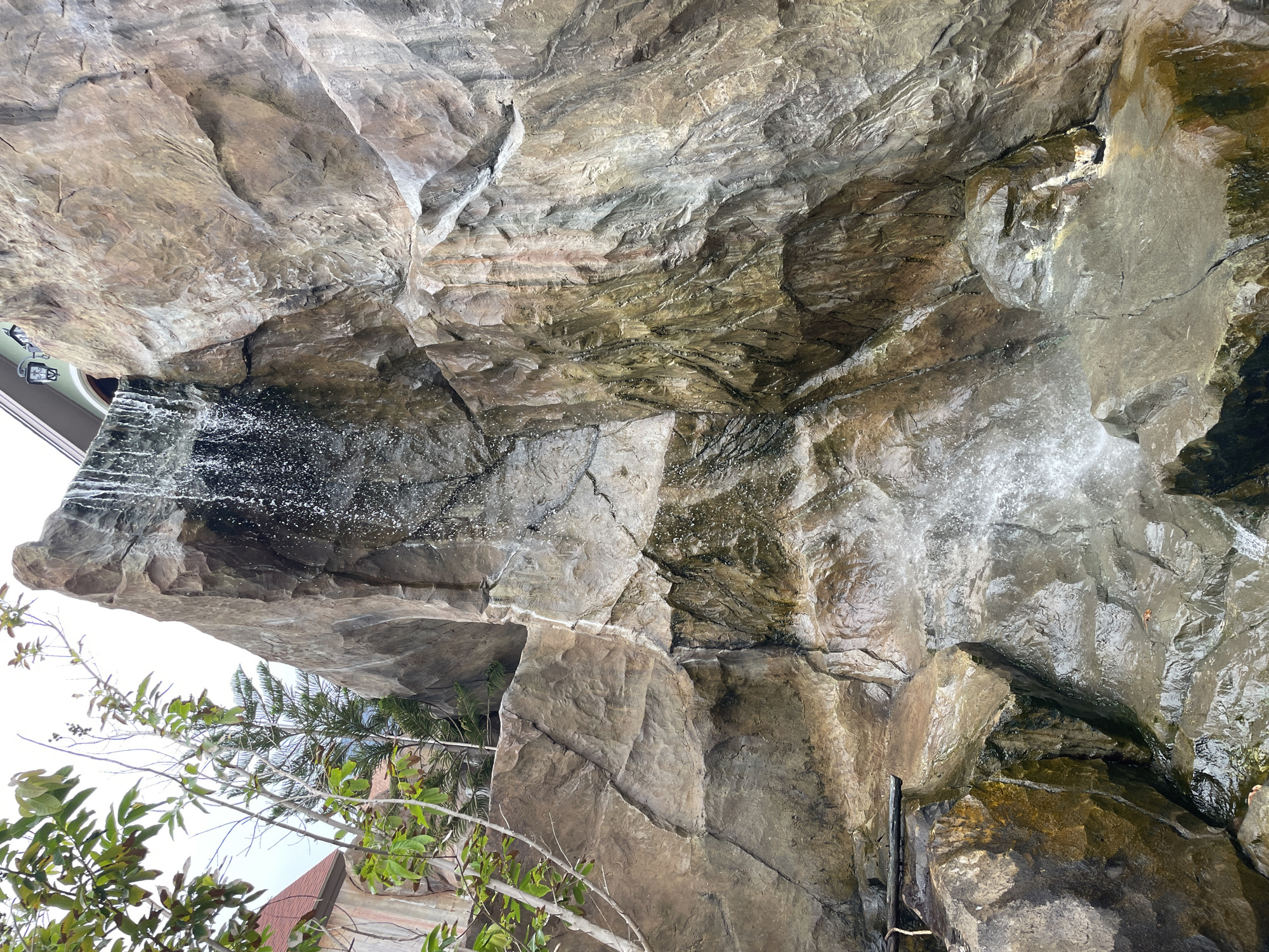 This is a vertical, medium-distance, eye-level shot of a man-made rock formation with a waterfall. The rock is textured and appears to be made of concrete or a similar material designed to mimic natural stone. Water cascades down the rock face from the top right, creating a spray and pooling at the bottom on various rock ledges and in a pool of water. The environment is lush, with green foliage visible on the left side of the frame, and a building with a red roof and light green walls can be seen peeking out from behind the foliage on the far left. The scene is likely located in a resort or theme park setting, possibly at the Pattaya Floating Market in Pattaya City, Thailand, given the architectural style of the building. There are no people or animals visible in the image. The lighting suggests it is daytime, with diffused light.