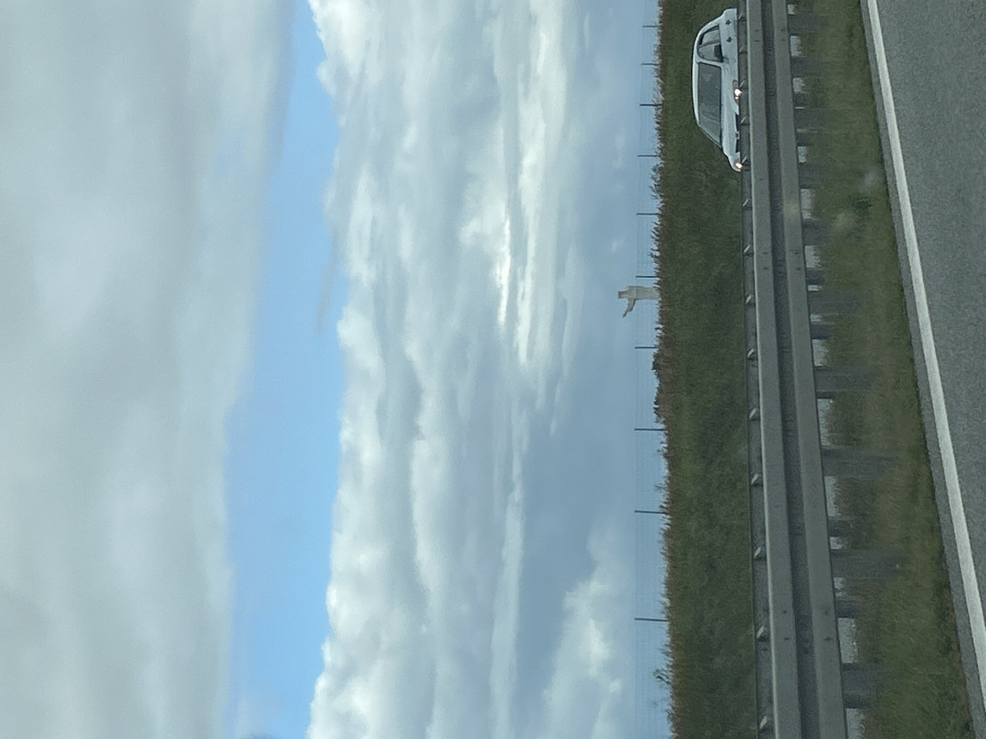 The image shows a wide shot of a highway in Lubinicko, Poland, with a statue in the distance. A white car is driving on the highway in the foreground, and a statue of what appears to be Jesus Christ is visible on a hill in the background. The sky is cloudy, with patches of blue visible. The overall scene suggests a typical day on the road in a rural area.