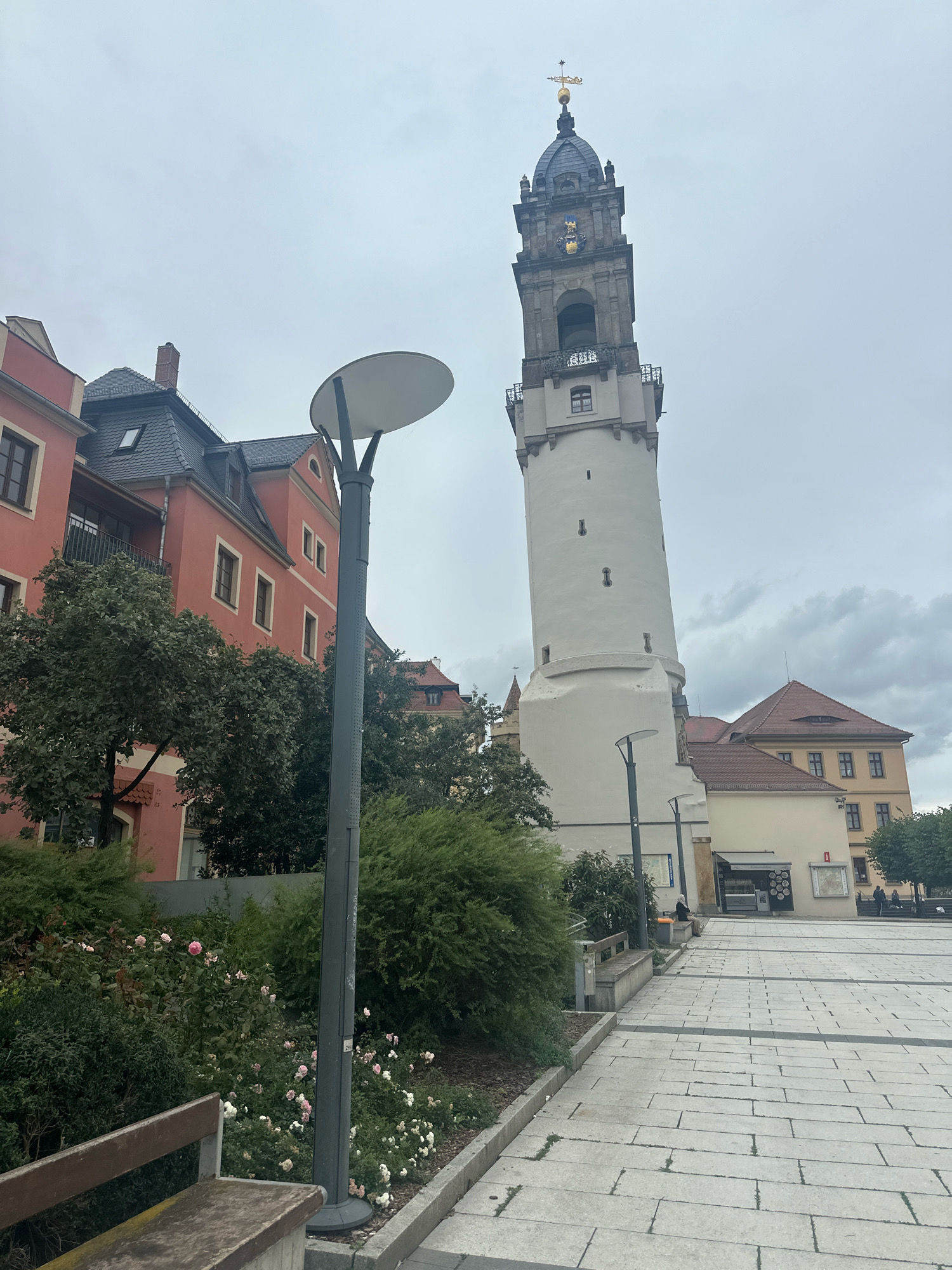 The image shows a view of the Ortenburg Castle in Bautzen, Germany, on an overcast day. In the foreground, a paved plaza with a gray tiled surface extends into the distance. To the left, a bench is visible next to a garden bed with pink and white roses and green bushes. A tall, gray lamppost with a circular light fixture stands in the middle ground, between the garden and the plaza.
Dominating the background is the striking white tower of the Ortenburg Castle, a prominent landmark. The tower has a round base that tapers upwards, and is topped with a dark, multi-tiered spire featuring a weather vane. Several windows are visible on the tower. To the right of the tower, a cluster of buildings with red tiled roofs and light-colored facades can be seen. One building has a prominent clock face. Other buildings with lighter facades and red roofs are visible further to the right.
In the mid-ground, several trees with lush green foliage are scattered throughout the scene, particularly on the left side near the pink building. A person is seated on a bench in the far distance on the right side of the plaza, facing away from the camera. Another person appears to be walking in the distance to the right. The overall atmosphere is subdued due to the cloudy sky.