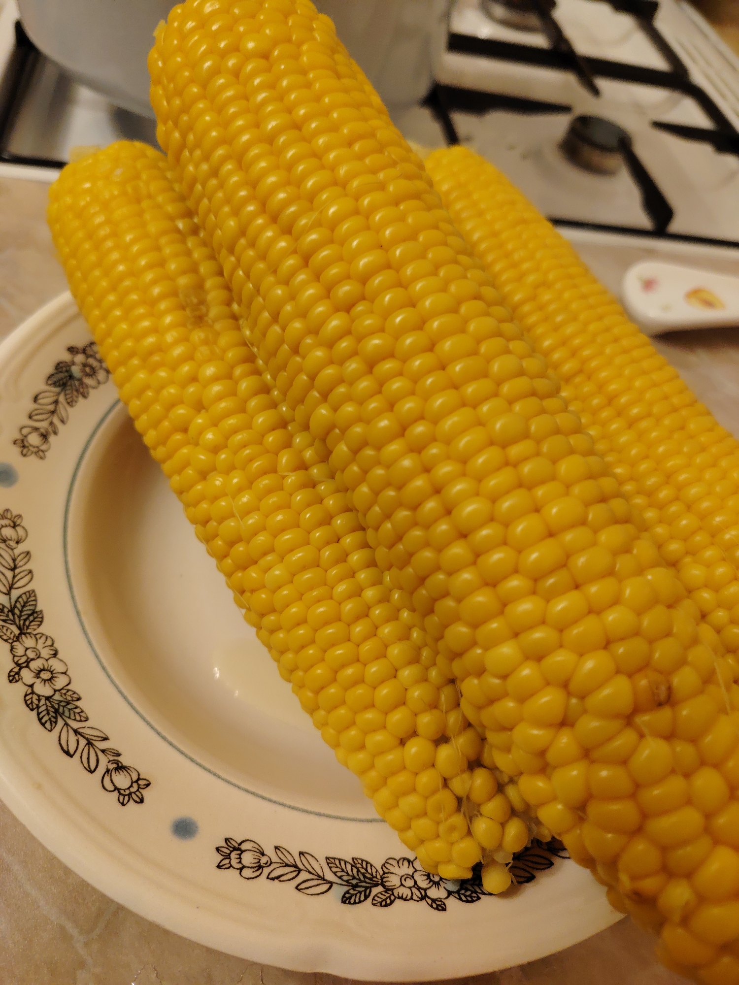 A close-up, slightly overhead shot shows three ears of cooked corn resting on a white plate with a floral pattern. The corn is bright yellow and appears to be freshly cooked. The plate has a blue rim and a black floral design with small blue dots scattered around the edge. The background is blurred, but shows a stovetop with burners and a white pot. The scene is indoors, likely in a kitchen. There are no people visible in the image. The location is identified as Konakovo, Russia.