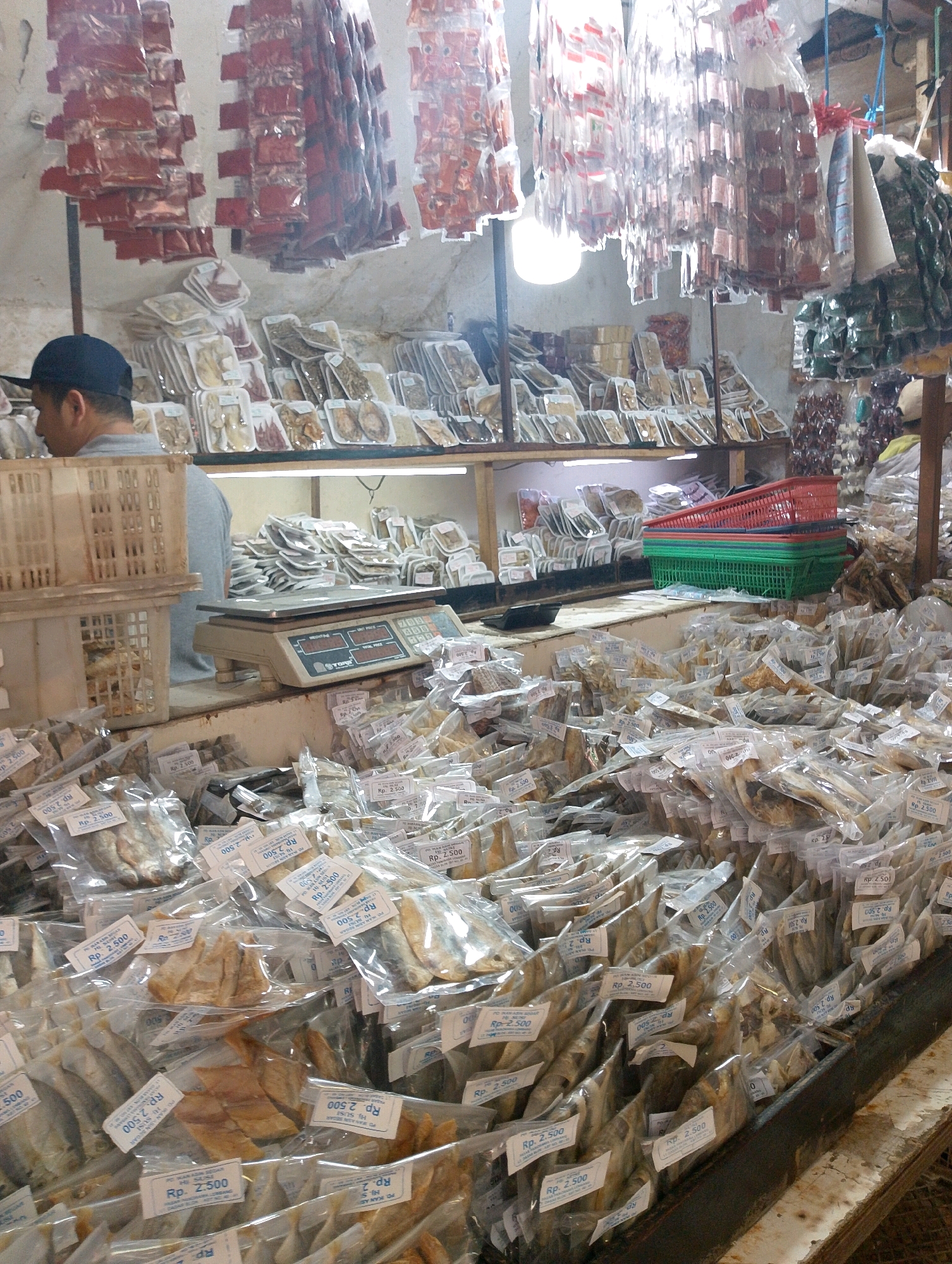The image depicts an indoor market stall in Lembang, Indonesia, filled with a variety of dried fish products for sale. In the foreground, a large quantity of individually packaged dried fish are neatly arranged, many with price tags labeled "Rp. 2.500". The packaging appears to be clear plastic, allowing the contents to be visible. Behind this display, there is a counter with a digital scale and a person wearing a black cap and a grey shirt, partially visible from the side. Further in the background, shelves are stocked with more packaged food items, some in translucent wrappers and others in darker, possibly red, packaging. Suspended from the ceiling are rows of what appear to be dried chili products or other condiments, also packaged individually. Several plastic crates and stacks of colorful plastic baskets (red and green) are visible on a shelf. The overall impression is of a busy market environment with an abundant display of food goods.