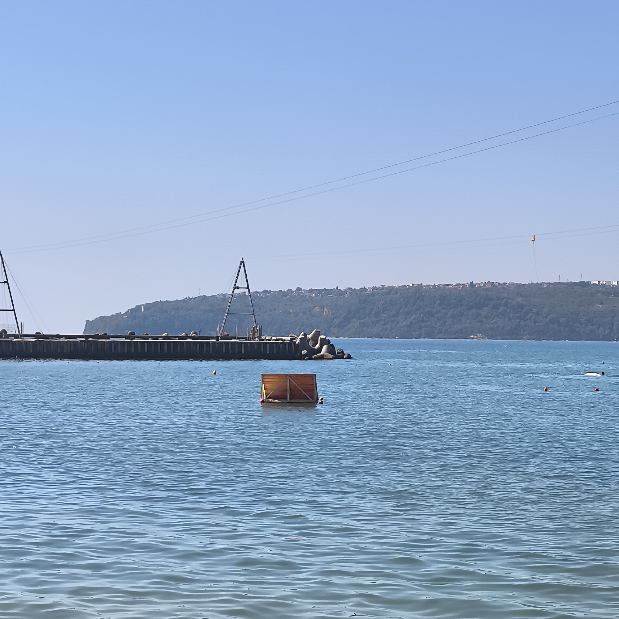 The image depicts a view of the Varna Bay in Varna, Bulgaria, on a clear, sunny day. In the foreground, the calm blue water shows gentle ripples. A wooden, floating structure with a rectangular frame is positioned in the middle of the water, with a red buoy attached to its side. Further out, a long pier extends into the bay, supported by sturdy pilings and adorned with large, conical concrete wavebreakers at its end. Two prominent A-frame structures are visible on the pier, suggesting some form of construction or support for overhead cables. In the background, a lush green, tree-covered hillside rises, dotted with buildings and houses of a settlement. Overhead, thin, dark lines representing power lines or cables stretch across the clear blue sky. The scene conveys a peaceful, daytime atmosphere by the sea, with a mix of industrial and natural elements.