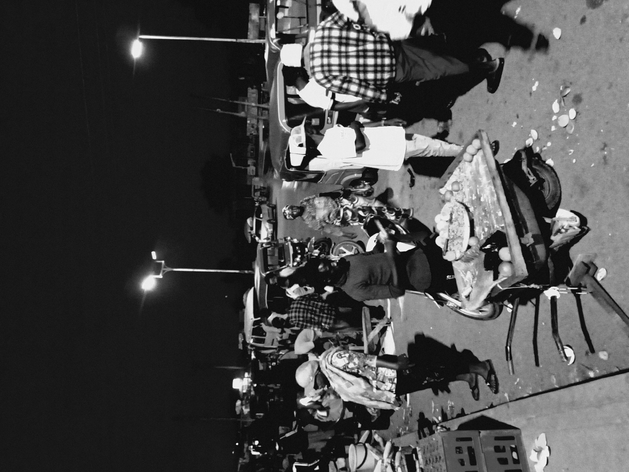 This black and white, grainy image captures a bustling night street scene, likely in Bukuru, Nigeria. The photograph is oriented sideways, with the main activity unfolding vertically across the frame.
**Subjects:** Numerous people are gathered along what appears to be a busy roadside. Towards the upper center, a person wearing a distinct plaid shirt is seated on a scooter or motorcycle, facing towards the right. Nearby, another individual stands next to a street vendor's cart or table. Several other people are visible in various positions, some near parked motorcycles, others standing.
**Objects:** The scene is populated by multiple scooters or small motorcycles, on which some individuals are seated. A central feature on the lower right is a vendor's makeshift table or cart, laden with several spherical items, possibly fruits or other food products, and other small goods. Bright streetlights are visible on the left side of the frame, emitting strong light that illuminates parts of the street and casts distinct shadows. On the ground, particularly around the vendor's setup, small, circular objects (which could be coins or discarded debris) are scattered. A dark crate or basket is also visible near the bottom right corner.
**Scene/Setting:** The setting is an active urban or semi-urban street at night, confirmed by the dark background and the reliance on artificial lighting from the streetlights. The ground appears to be paved, suggesting a developed area. The overall atmosphere conveys a sense of everyday life and commerce taking place after dark.
**Activity or event taking place:** The primary activity revolves around street vending and informal commerce. People are congregating, seemingly interacting with the vendor, possibly buying or selling goods. The presence of multiple motorcycles suggests transport and a dynamic, active environment. The scene depicts a common sight of social gathering and economic exchange in a public space during the evening hours.
**Notable details:** The image's black-and-white presentation and noticeable graininess contribute to a candid, documentary aesthetic. The strong contrast between the bright streetlights and the dark surroundings effectively highlights the figures and objects in the foreground. There is no visible text discernible within the image.