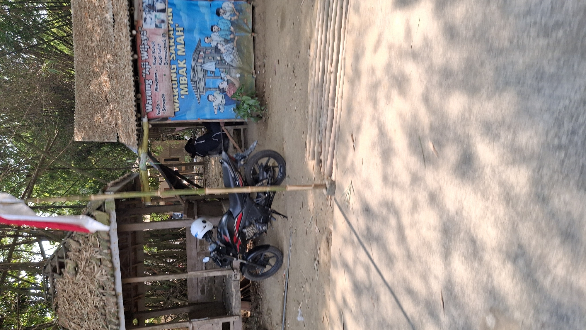 The image captures a roadside scene in Winong, Indonesia, featuring a small outdoor eatery and a parked motorcycle. The eatery has a rustic appearance with a thatched roof and wooden structure. A blue banner with text in Indonesian is prominently displayed, advertising "Warung 'Aji Wijaya'" and "WARUNG SAKAPAHAN 'MBAK MAH'". The banner also includes illustrations of people.

In front of the eatery, a black motorcycle with a white helmet resting on its seat is parked. To the left of the motorcycle is a simple wooden stall with a thatched roof. The ground is unpaved and appears to be dirt and sand, with some scattered dry leaves. A bamboo pole stands vertically in the foreground, partially obscuring the scene. In the background, dense bamboo foliage creates a natural, verdant backdrop. The overall impression is that of a humble, locally run establishment in a rural or semi-rural setting.