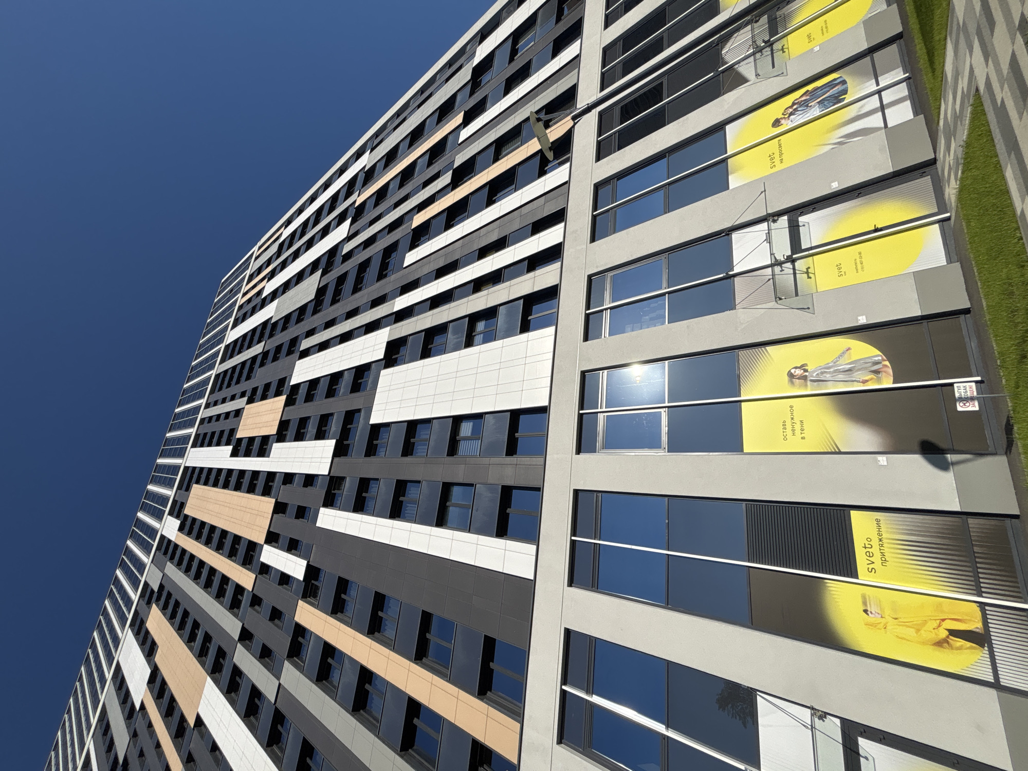 A low-angle, upward shot reveals a high-rise building with a varied color palette, juxtaposed against a clear blue sky. The building's exterior incorporates black, white, beige, and gray panels, creating a striped effect across its facade. Rows of windows, reflecting the sky, are interspersed throughout.
On the right side of the building, large advertisements, predominantly yellow with black text, are affixed to the structure. The text appears to be in Russian and includes the words "sveto," "притяжение," and "оставь ненужное в тени." There is also a warning sign prohibiting dogs. One of the ads features an image of a person in a yellow coat.
The location is likely Vsevolozhsk, Russia, based on the Cyrillic text in the advertisements. The environment is urban, suggested by the architectural style and the presence of advertisements.