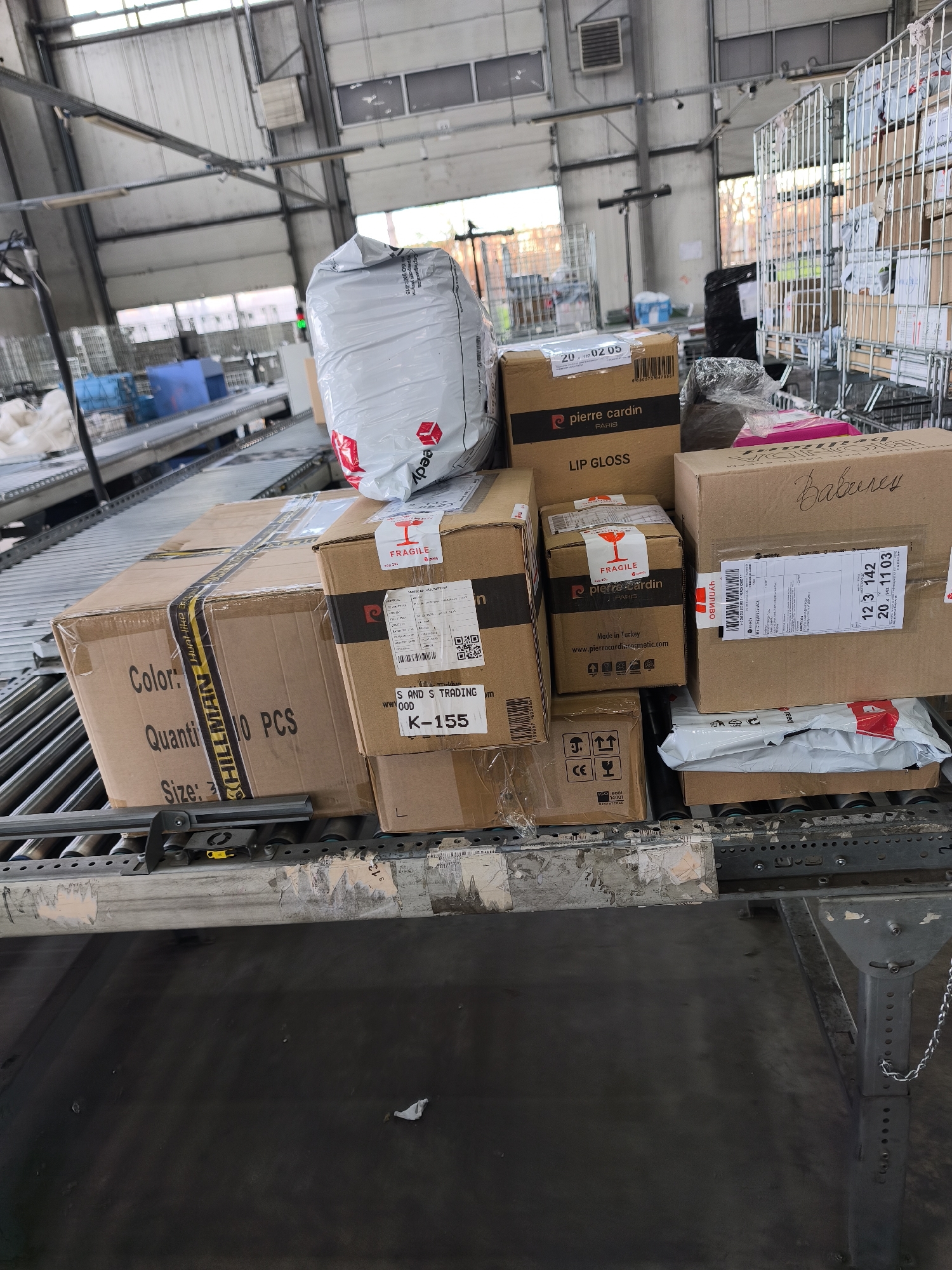 This image depicts an indoor scene within what appears to be a logistics or postal sorting facility in Burgas, Bulgaria. A long conveyor belt system dominates the foreground and midground, laden with various packages and parcels, indicating an ongoing or recently paused operation of shipping and handling.
The primary subjects are numerous cardboard boxes and one large white plastic bag, all destined for or moving through the distribution process. The packages are diverse in size and markings. Notable objects include the metal framework of the conveyor system, metal cages used for storage or transport in the background, and industrial lighting fixtures on the high ceiling. No people are visible in the frame.
Notable details include the commercial nature of the packages. Several boxes feature the "pierre cardin PARIS" brand, with one explicitly labeled "LIP GLOSS" and another indicating "Made in Turkey". Another box mentions "S AND S TRADING" and "K-155". A larger box has "Color:", "Quanti 10 PCS", and "Size: 3". Many boxes display "FRAGILE" stickers, some with the broken glass symbol, along with handling arrows, a CE mark, and an ISO 9001 certification. The white plastic bag, potentially a bulk shipment, has a red logo and the word "eedy". Bulgarian text is visible on some labels, including "Чупливо" (Fragile) and "Вавилен". Shipping codes like "20,702 05", "12 3 142", and "201103" are also present on various labels. The setting is clearly an industrial environment, characterized by concrete floors, metal structures, and an efficient system for moving goods. The lighting is artificial, suggesting the image could have been taken at any time of day.