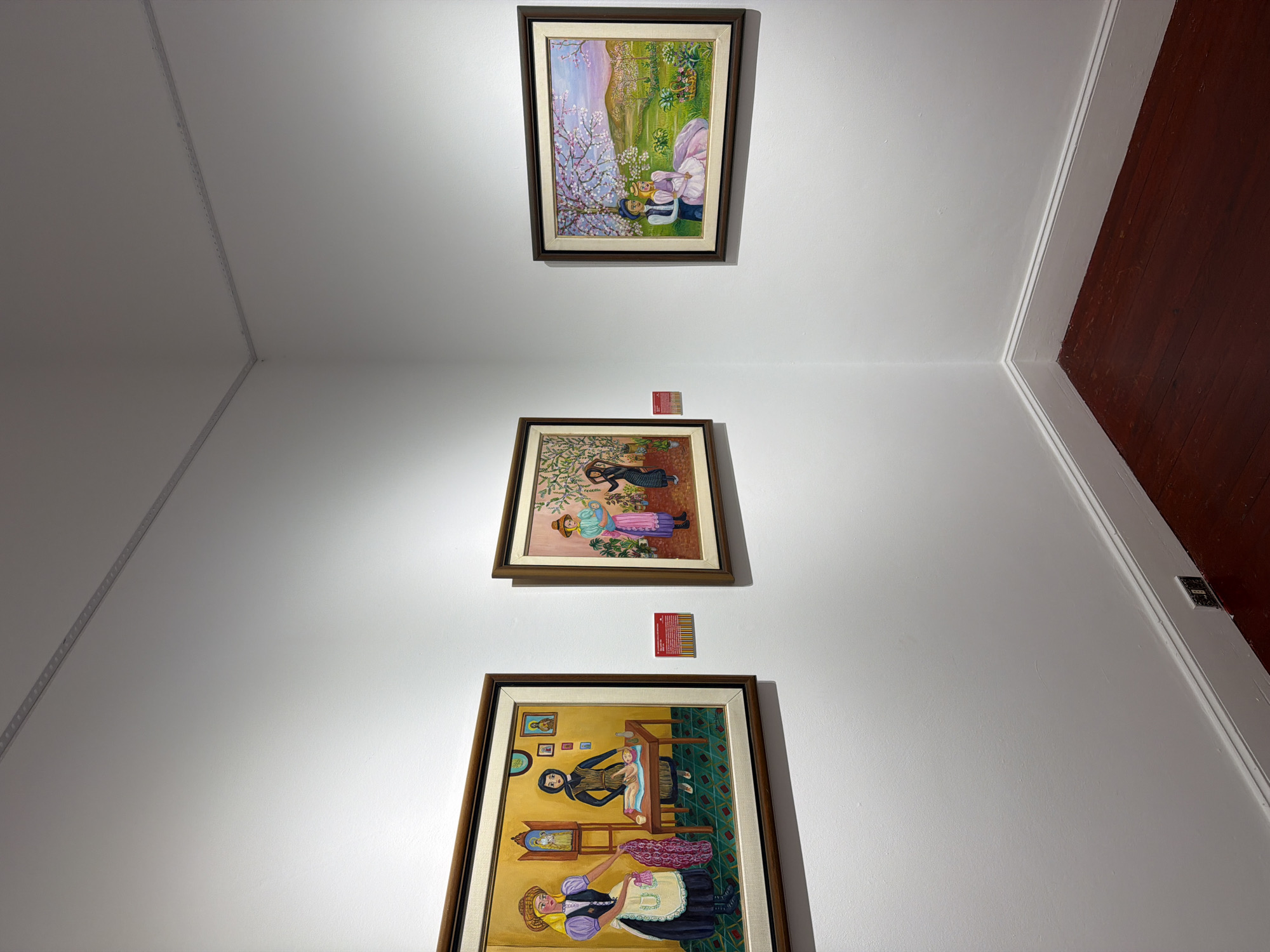 Three framed paintings are displayed on a white wall in a corner of a room with dark red hardwood floors. The painting on the left depicts two women in traditional attire in an indoor setting, with one woman tending to a baby on a table. The center painting shows two women in a garden with patterned wallpaper. The painting on the right portrays a couple sitting in a blooming tree-lined landscape. Red labels are attached to the wall below the left and center paintings. A light fixture runs along the ceiling in the corner.