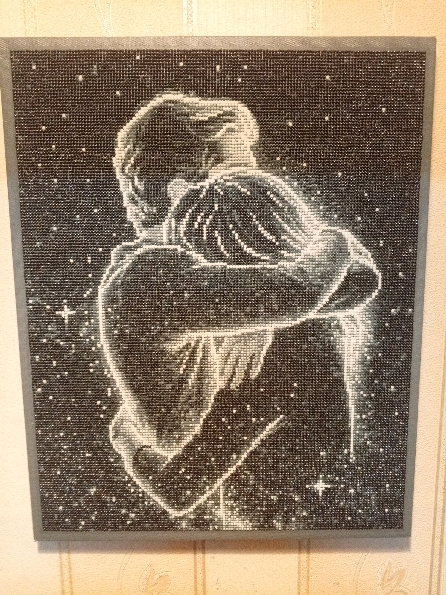 A framed, square diamond art piece depicting two figures embracing against a dark background is mounted on a light-colored, patterned wall. The figures are predominantly white, formed by the diamond beads, with a diffuse, luminous quality that contrasts with the solid black background filled with interspersed white dots resembling stars. The figures appear to be a man and a woman, with the man enveloping the woman in a hug. The surrounding frame is gray. The location is not depicted, only the object on the wall is visible.