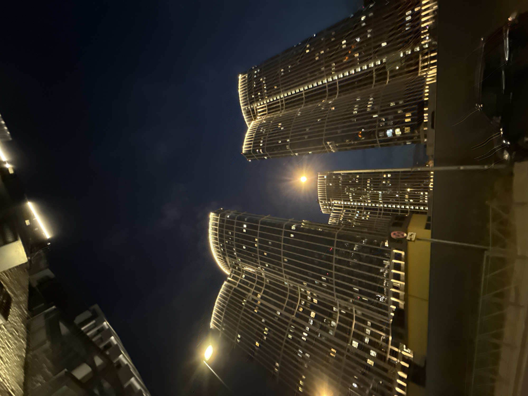 Captured at night, the image showcases the cityscape of Khimki, Russia. The focal point features two tall, modern buildings illuminated with horizontal lines of lights, revealing numerous windows across their facades. A street lamp casts a bright glow from below, lighting up the base of the structures. In the background, another building, partially visible, echoes the architectural style of the main buildings. The surrounding environment is dark, emphasizing the illuminated structures against the night sky. The angle suggests the photo was taken looking upwards, capturing the scale and height of the buildings. The photo also show a part of the reflection in what seems to be the window or glass structure, this is not the main element in the image and not necessary to describe it.
