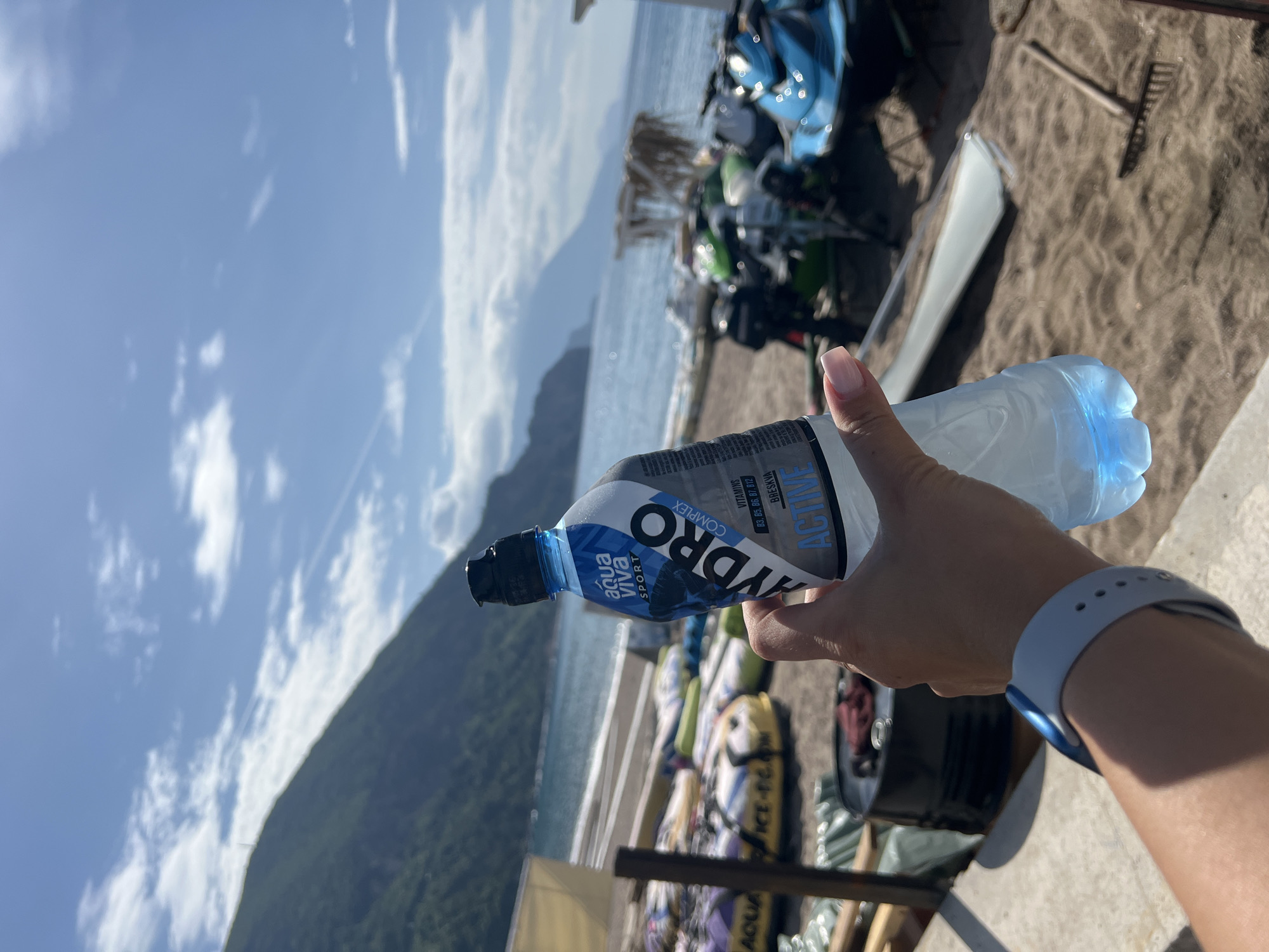 A person holds a bottle of Aqua Viva Hydro Sport water against a backdrop of a beach near Prijevor, Montenegro. The bottle, predominantly gray and white with blue accents, features text including "HYDRO COMPLEX" and vitamin information. The person's hand and part of their arm are visible, adorned with a light blue watch. Behind the bottle, the scene reveals a beach with various water sports equipment and vehicles. The background also showcases the Adriatic Sea, a mountainous landscape, and a partly cloudy sky.