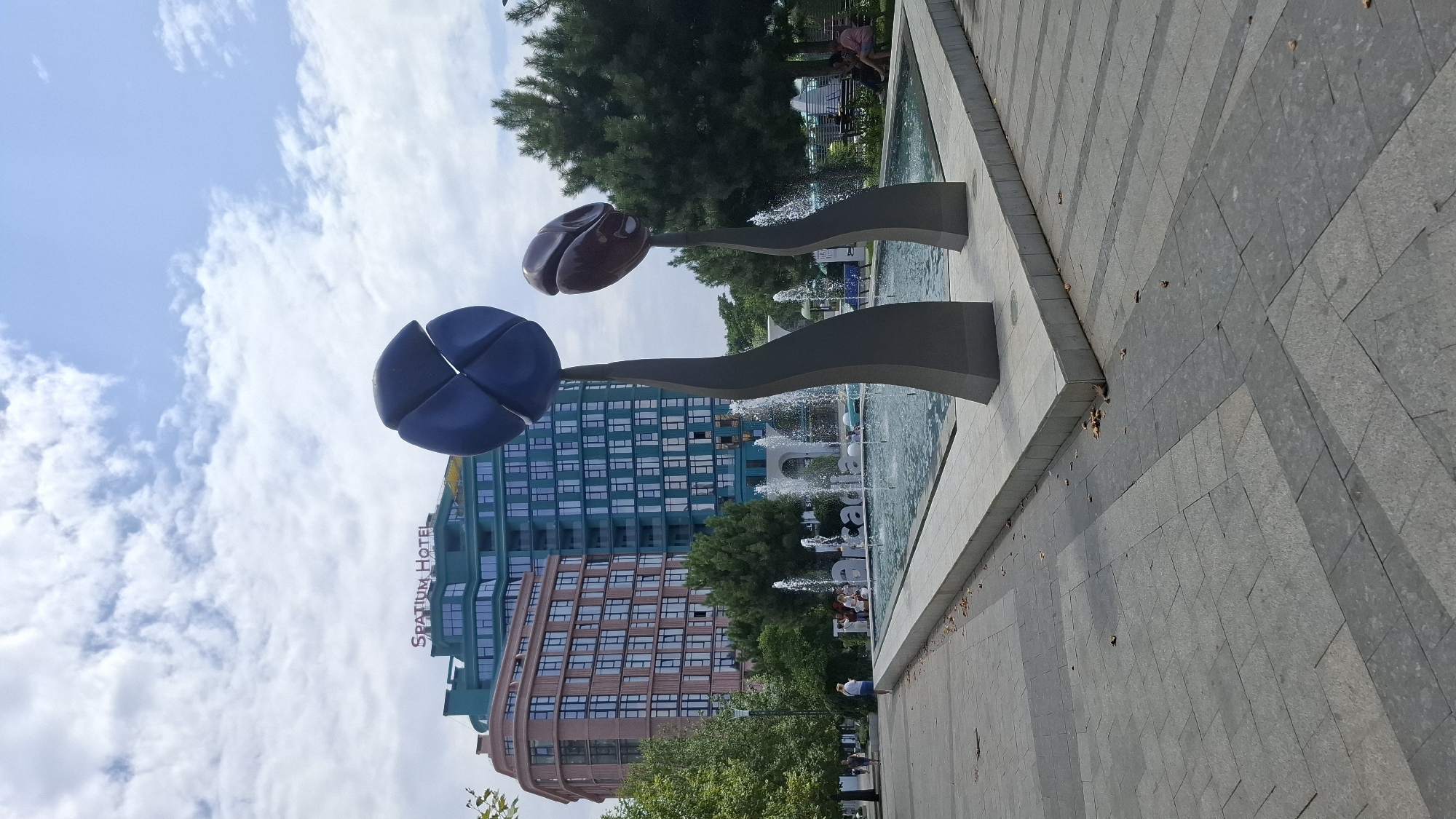 In a sunlit outdoor plaza, a wide-angle shot captures a modern urban scene with a focal point on a decorative fountain and accompanying sculptures. Two abstract sculptures, resembling large stylized flower buds or seeds, rise from the ground, their dark grey bases tapering upwards to support dark blue and reddish-brown rounded forms.
The fountain itself is a rectangular pool with water jets creating a misty spray. In the background, two contemporary buildings dominate the skyline. The taller building on the right is predominantly teal with numerous windows, and it features a prominent red sign at the top that reads "SPATIUM HOTEL." To its left is a slightly shorter building with a reddish-brown facade, also characterized by a grid of windows.
Surrounding the fountain and plaza area are green trees, providing a natural contrast to the architecture. Several people are visible in the scene: a few individuals walk in the distance on a paved path to the left, and a small group is seated near the fountain. On the far right, two individuals are seated on a bench. The ground of the plaza is paved with large, rectangular grey tiles arranged in a geometric pattern. The sky above is a bright blue with scattered white clouds.