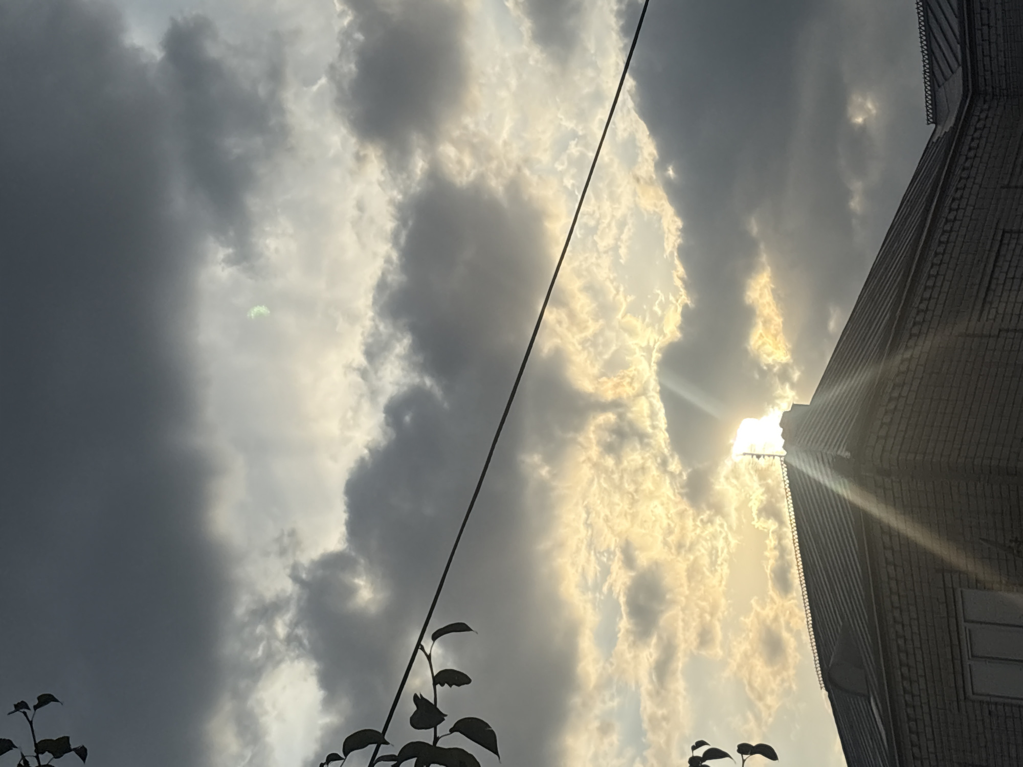 The image shows a cloudy sky over a brick building in Georgiyevsk, Russia. The sun is partially obscured by dark clouds, with rays of light breaking through and casting a bright glow on the clouds. A black wire runs diagonally across the upper portion of the image, with dark leaves from a tree branch visible in the upper left corner. The building appears to be a residential or commercial structure with a dark, sloped roof and visible brickwork. The overall mood is dramatic, with the interplay of light and shadow suggesting an approaching storm or a change in weather.