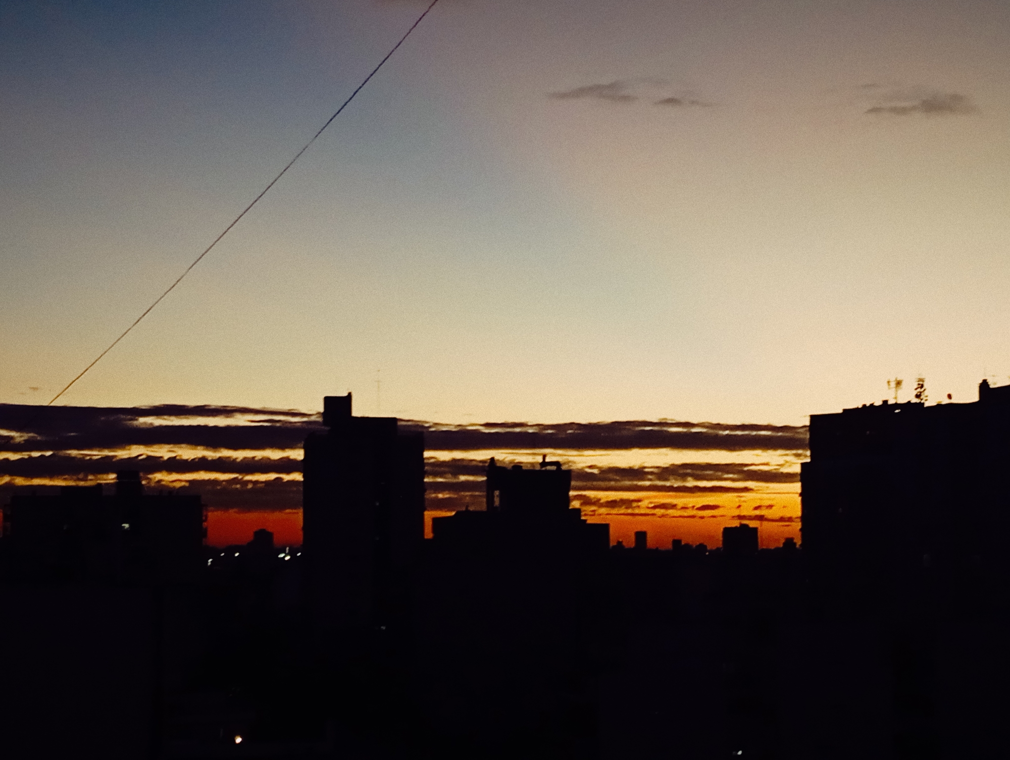 The image depicts a cityscape at sunset in Buenos Aires, Argentina. Silhouetted buildings dominate the foreground, with visible rooftops and structures. The sky transitions from blue to orange, with dark, horizontal cloud bands near the horizon. A powerline is visible in the upper left corner.