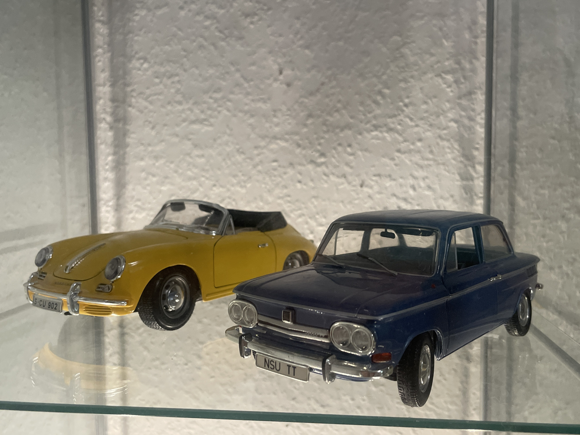 The image displays two toy cars on a glass shelf. The car in the foreground is a dark blue sedan with the license plate "NSU TT". To its left and slightly behind is a yellow convertible with the license plate "S-CU 902". The background is a textured white wall. There is no indication of Iserlohn, Germany in the image.