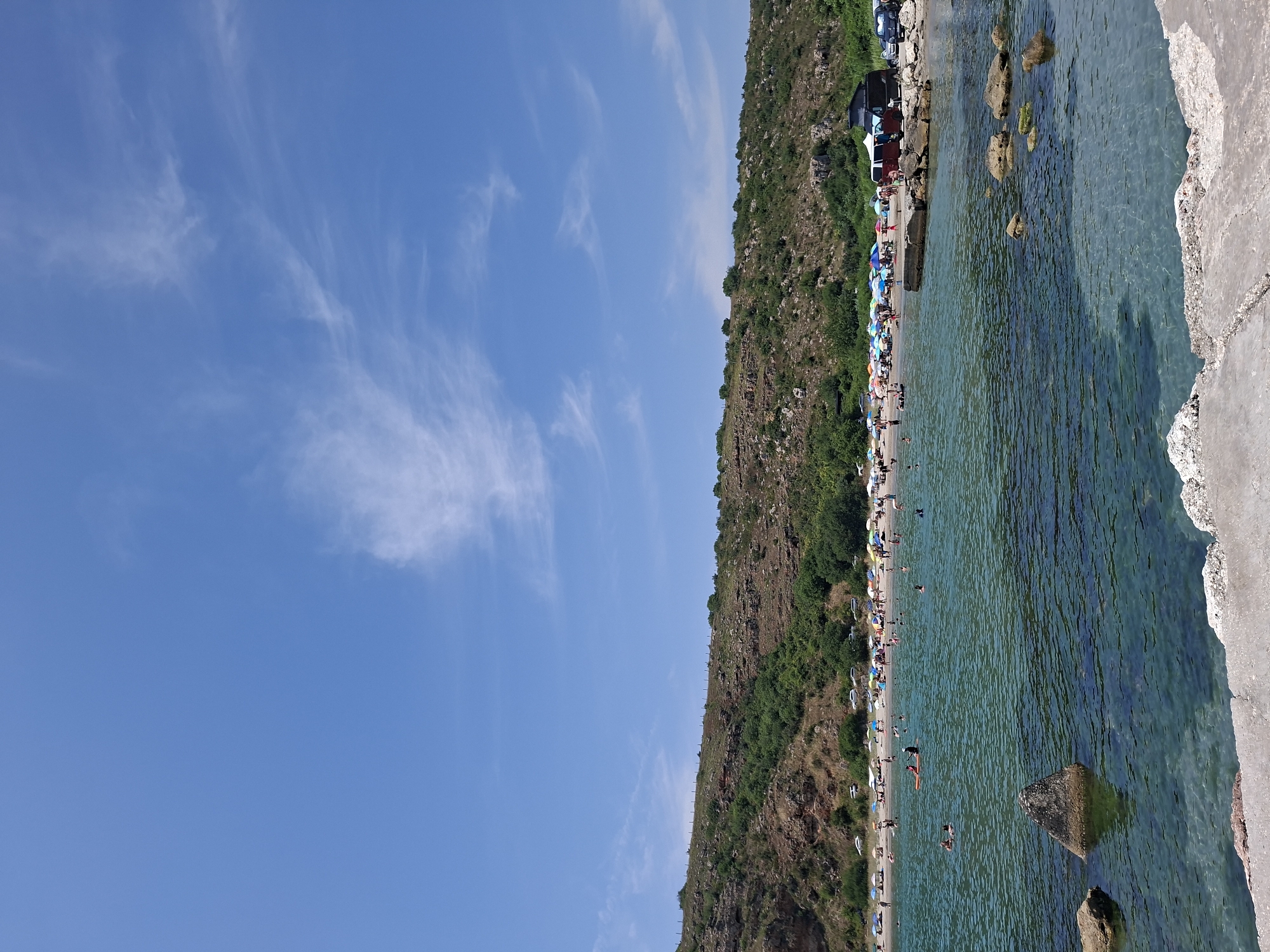 This is a medium shot, eye-level, outdoor photograph of a crowded beach on a sunny day, located in Balgarevo, Bulgaria. The scene captures a panoramic view of a crescent-shaped beach nestled against a rocky, vegetated hillside. The water in the foreground is clear and turquoise, with visible rocks and a concrete pier in the bottom right corner. The middle ground shows the beach populated with numerous sunbathers, umbrellas, and a few cars parked along the edge. People are seen swimming, walking, and relaxing on the sand. The background features the sloping hillside covered in green vegetation and scattered rocks, with a bright blue sky above adorned with wispy white clouds.