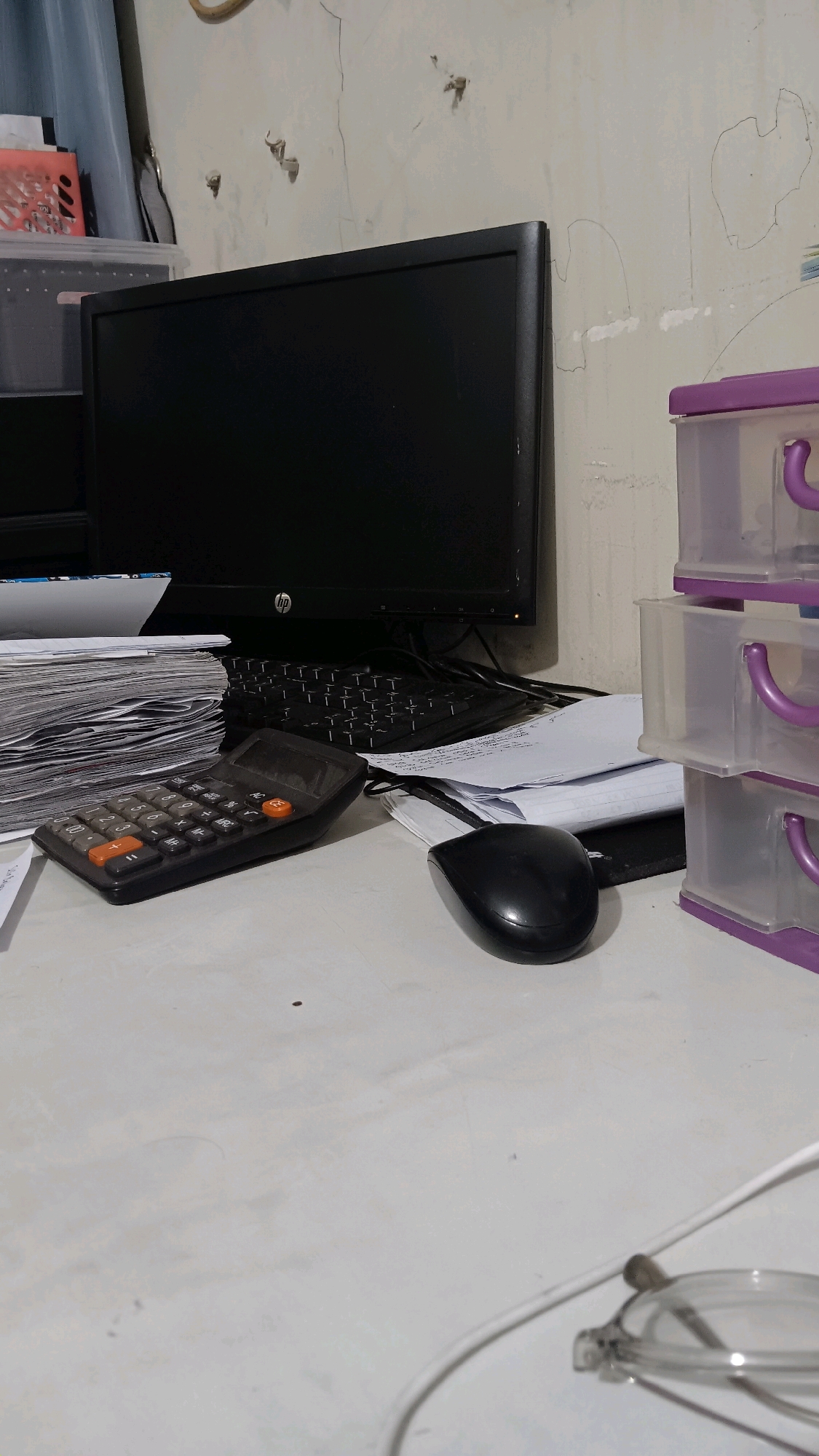 The image depicts a cluttered desk in what appears to be an office or study space. In the background, a black computer monitor is visible, with a cracked and textured wall behind it. To the left of the monitor, a stack of papers is partially obscured by a clear plastic container with a red-pink insert. In the foreground, a black calculator with orange and gray buttons sits on the desk, next to a black computer mouse. A stack of papers and documents is also present on the desk, some of which are unfolded. To the right of the monitor, a stack of clear plastic drawers with purple handles is visible. A white cord snakes across the desk towards the bottom right corner, where a pair of eyeglasses is partially visible. The overall impression is one of a work-in-progress or an actively used workspace. The location is identified as Pangebatan, Indonesia.