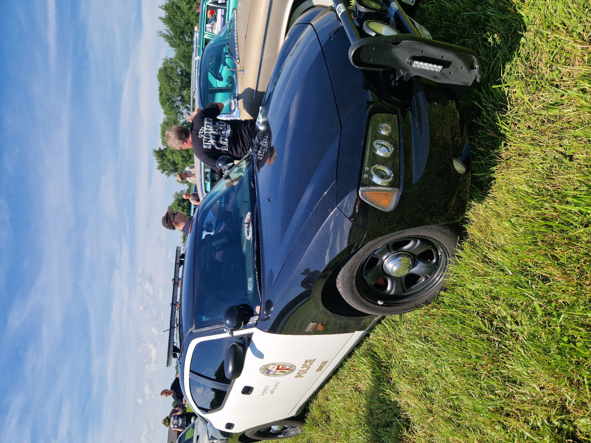 An exterior, eye-level, medium shot captures a black and white police car parked on a grassy field under a partly cloudy sky. The car, a Dodge Charger, is the primary subject. It has a prominent black push bar on the front, equipped with auxiliary lights. The car's side is marked with the word "POLICE" in gold lettering, along with a seal and the text "to protect and to serve." A person in a black t-shirt with a graphic design stands behind the car, facing away from the camera. Several other people are visible in the background, some standing near other vehicles. The environment appears to be an outdoor event or gathering, possibly a car show, given the presence of multiple vehicles and people. The ground is covered in green grass, and trees are visible in the distance. The sky is blue with scattered white clouds. The image is in sharp focus, capturing the details of the car and its surroundings. The location is identified as Edenberga, Sweden.