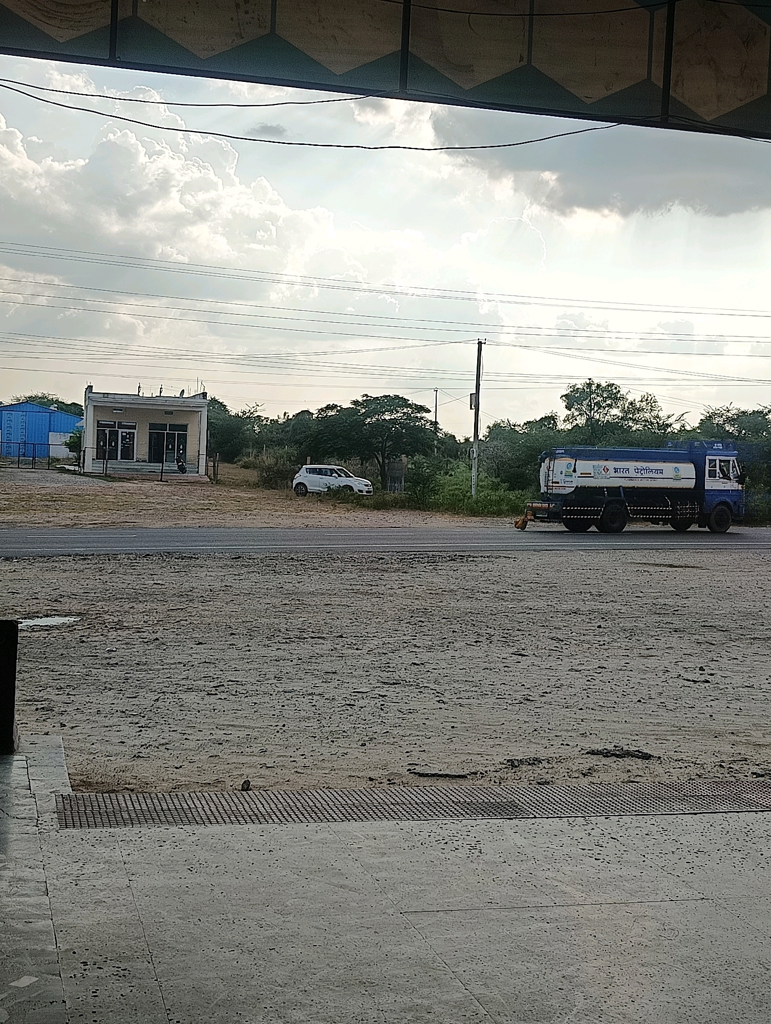 This is a medium-angle, vertical shot of a roadside scene in Chhapri Bari, India. The foreground is dominated by a rough, sandy, textured ground with a dark, metal grate. Behind this, a paved road runs horizontally across the frame, with a white car parked on the right side and a blue and white tanker truck labeled "Bharat Petroleum" driving on the road from left to right. In the background, there is a small, light-colored building with a dark roof and storefront windows, and behind that, a cluster of green trees. Overhead, dark power lines crisscross a cloudy sky.