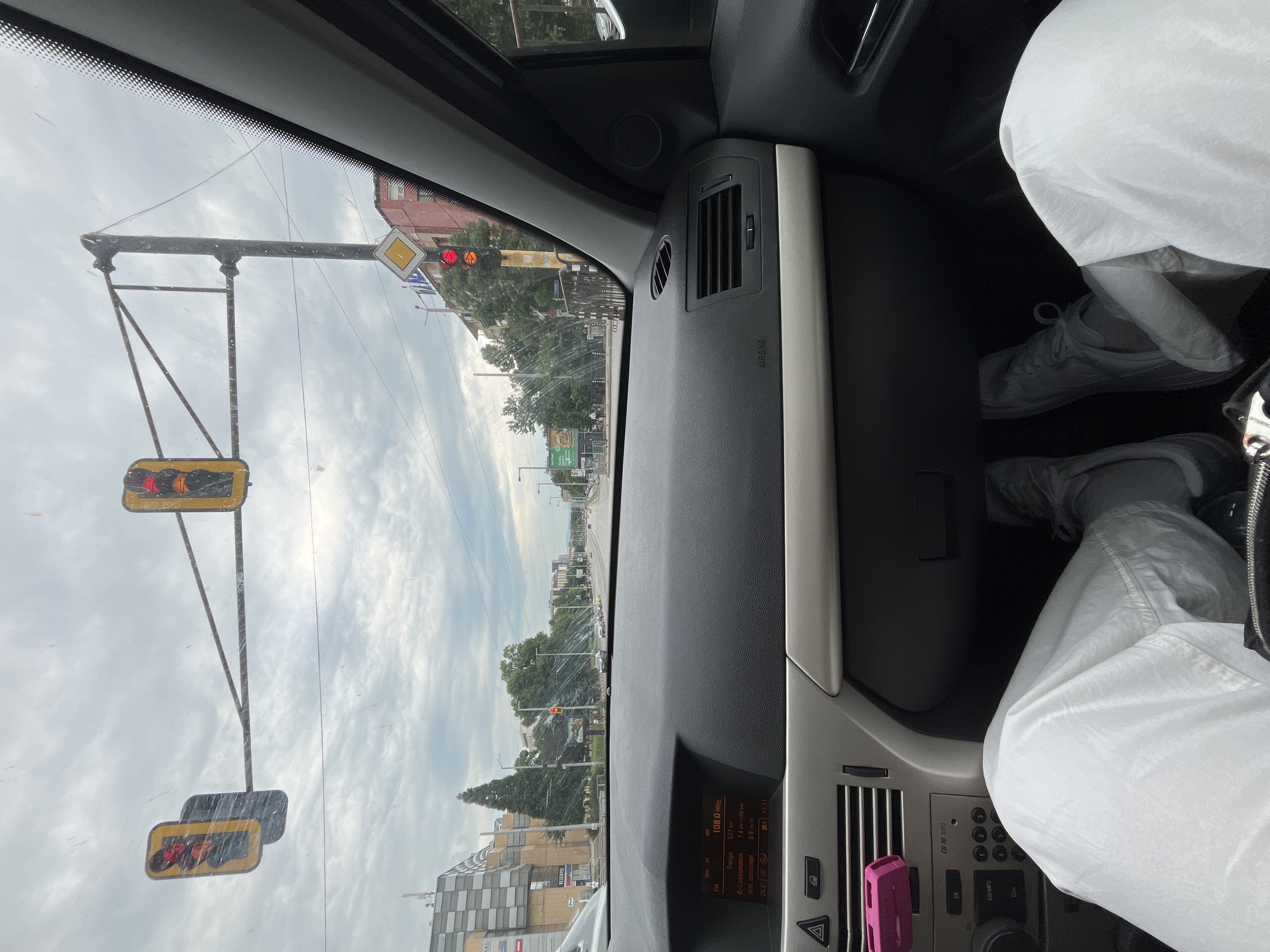 The image presents a first-person perspective from within the passenger compartment of an Opel Astra H automobile, looking forward through the windshield on an overcast day. The vehicle is stationary at a multi-lane urban intersection in Sofia, Bulgaria, identified by the distinctive architecture of the Serdika Center shopping mall visible on the left, specifically the building featuring a checkered black and white facade and signage for "RESERVED."
Overhead, two traffic lights displaying red signals are mounted on a dark metal gant. To the right, a vertical traffic light is also red, with a yellow diamond-shaped "priority road" sign positioned below it. The road ahead is asphalt, extending into the distance with several other vehicles visible. Trees and other urban foliage line the boulevard, separating the road from buildings. On the right side, an older, multi-story building of brick-like color is visible, along with a partially obscured green billboard advertising food.
Inside the car, the black dashboard with a silver-gray trim panel is prominent. The central digital display indicates "FM 108.0 MHz," "Range 637 km," "e-Consumption 7.4 l/100km," and "inst. consumpt. 0.9 l/h," along with "26.0 16." Below the display, a control panel with a radio (CD 50 MP3 capable) and climate buttons is visible. A pink USB charger is plugged into the leftmost air vent. The legs of a person, likely the passenger, are visible in the foreground, clad in white, loose-fitting trousers and light gray athletic shoes. The windshield shows numerous small spots, possibly watermarks or dust.